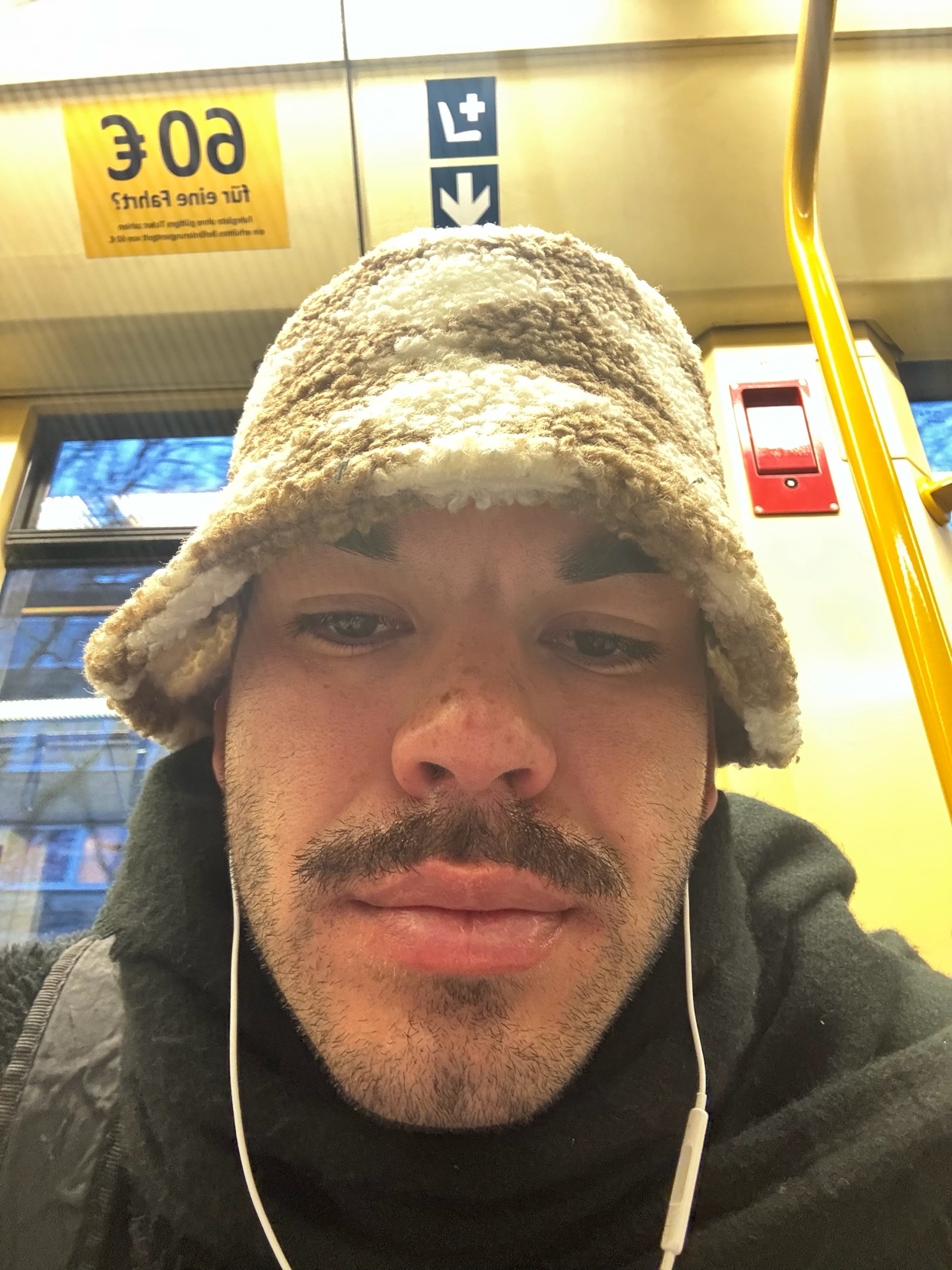 A man wearing a fuzzy bucket hat and earbuds takes a selfie on public transport.