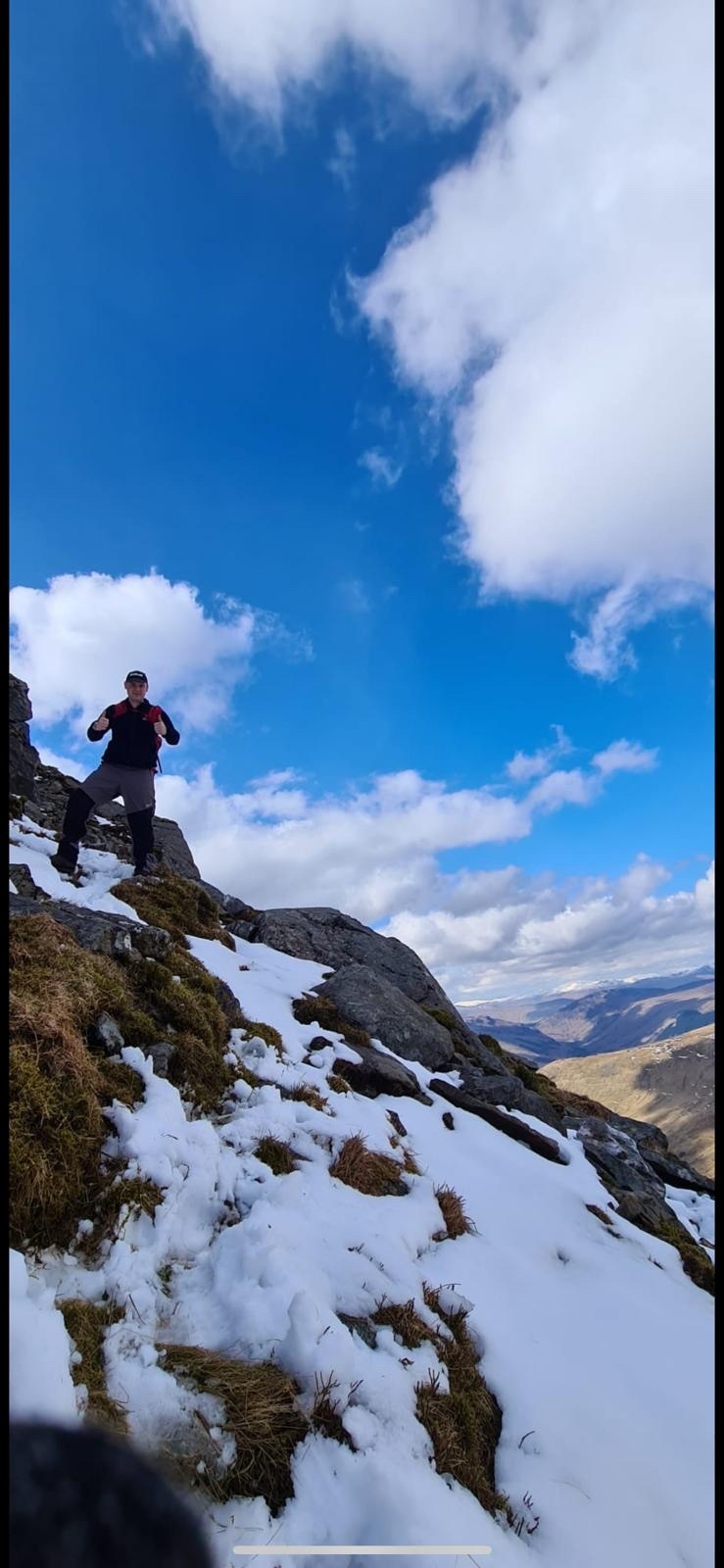 A man stands on a snowy, rocky mountain slope under a bright blue sky with fluffy clouds. The landscape stretches out into the distance, hinting at a vast, rugged terrain.