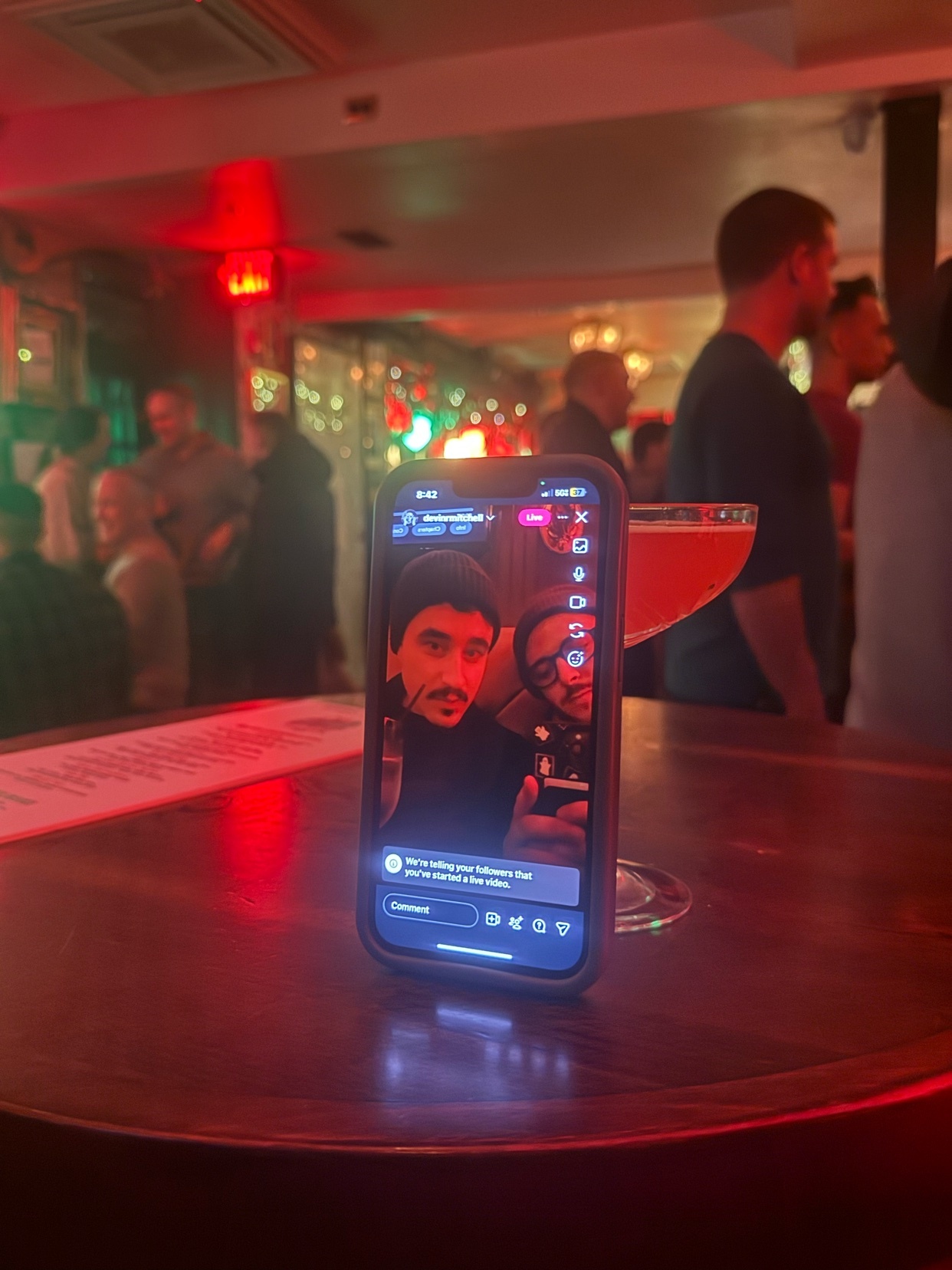A phone displays a live video of two men, set on a bar with a cocktail. The background shows a crowded bar scene.