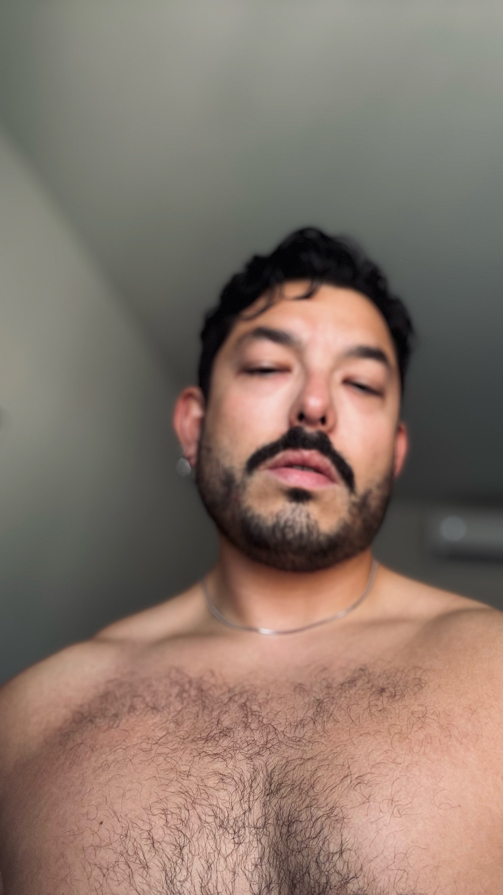 A man with dark hair and a beard poses shirtless with his eyes closed. The photo is a close-up portrait.