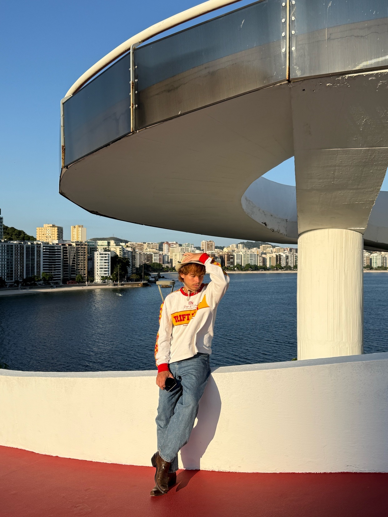 A man stands on a curved walkway overlooking a bay with a cityscape in the background. He is wearing a long-sleeved shirt, jeans, and a hat, with one hand resting on his head.