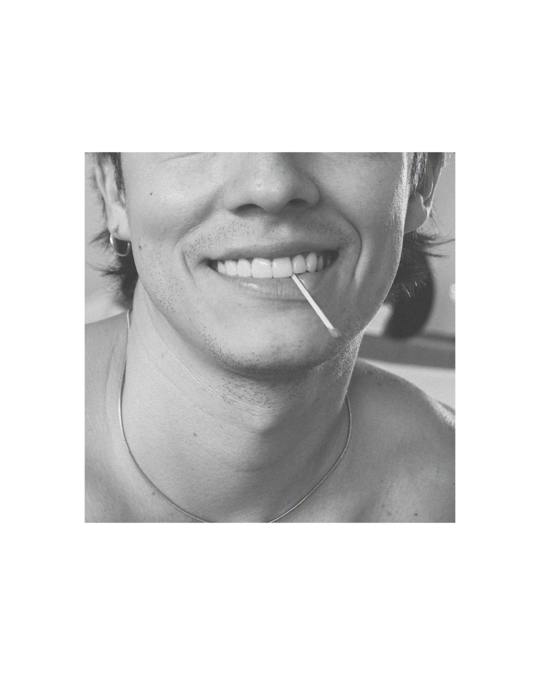 A black and white close-up photo shows a man smiling with a toothpick in his mouth. He is wearing a thin necklace.