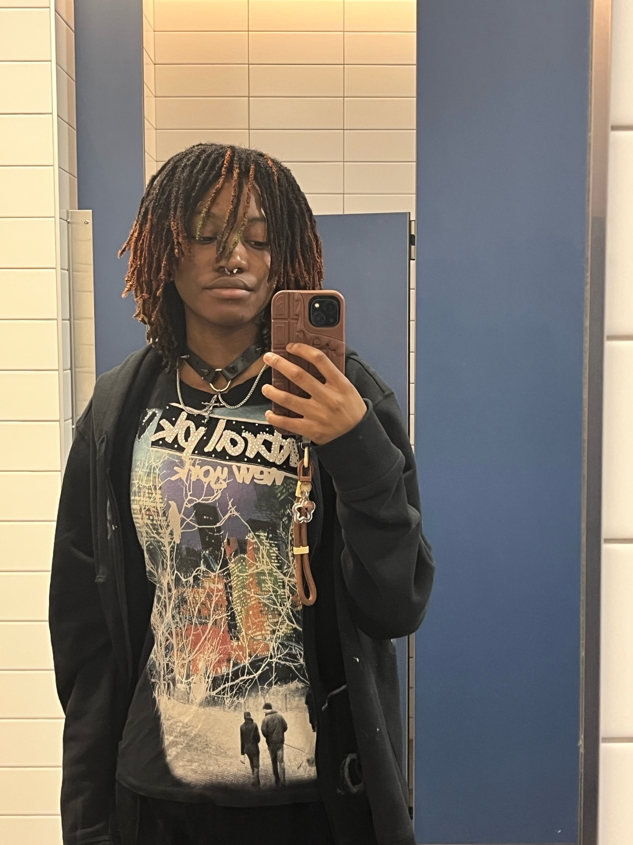 A young woman with dreadlocks takes a selfie in a bathroom mirror. She is wearing a graphic t-shirt, a black hoodie, and a choker necklace.