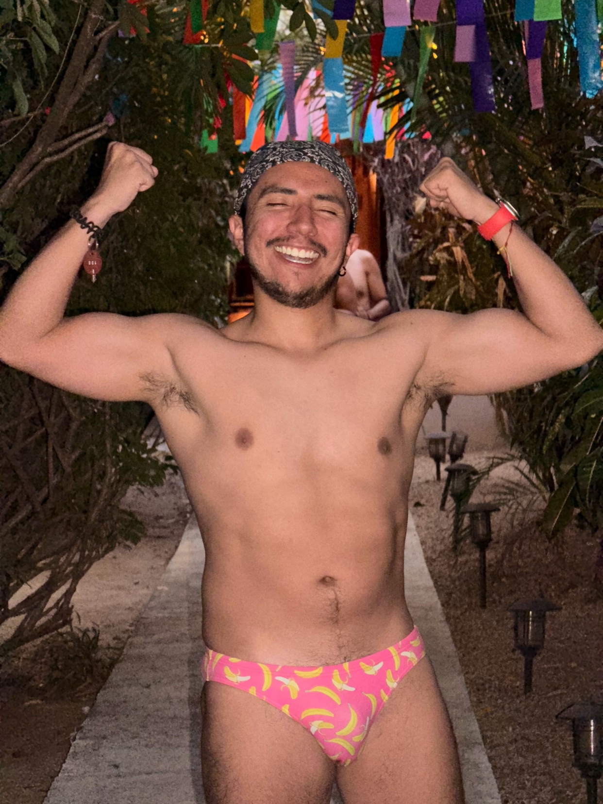A man with a bandana and a wide smile flexes his biceps while wearing pink banana-print swim briefs. He is standing outdoors on a path lined with small lights, with lush greenery and colorful flags in the background.