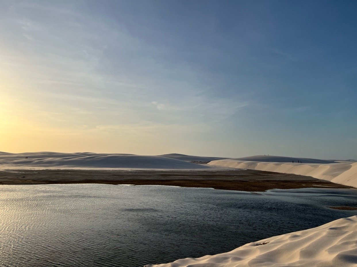 A stunning landscape of sand dunes and a lake under a clear blue sky. The golden hour light bathes the scene in a warm glow.