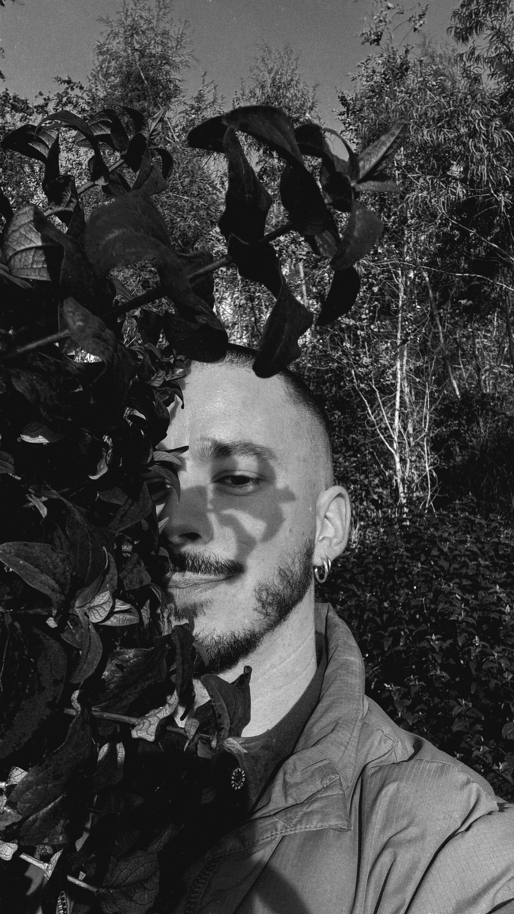 A black and white portrait of a man with a beard, partially obscured by leaves. The photo is taken outdoors.