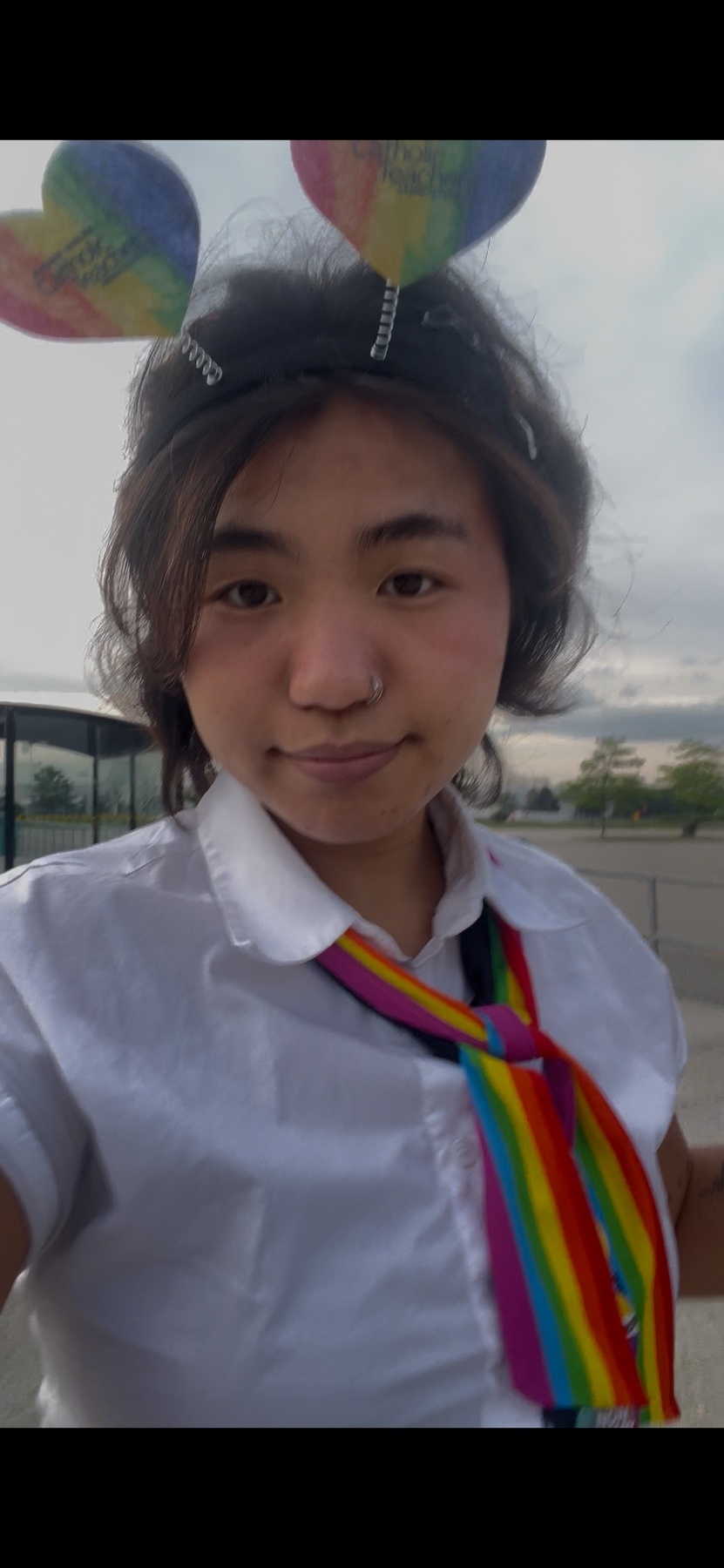 A young woman with dark hair and a nose ring smiles at the camera while wearing a white collared shirt and a rainbow-striped tie. She has a headband with two rainbow-colored hearts on it.