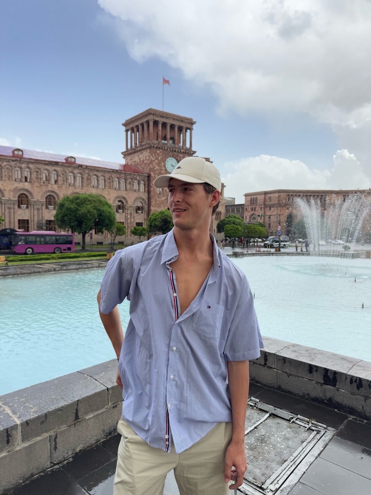 A young man in a light blue button-down shirt and beige shorts poses in front of a fountain and a large building with a clock tower. He is wearing a beige baseball cap and looking off to the side.