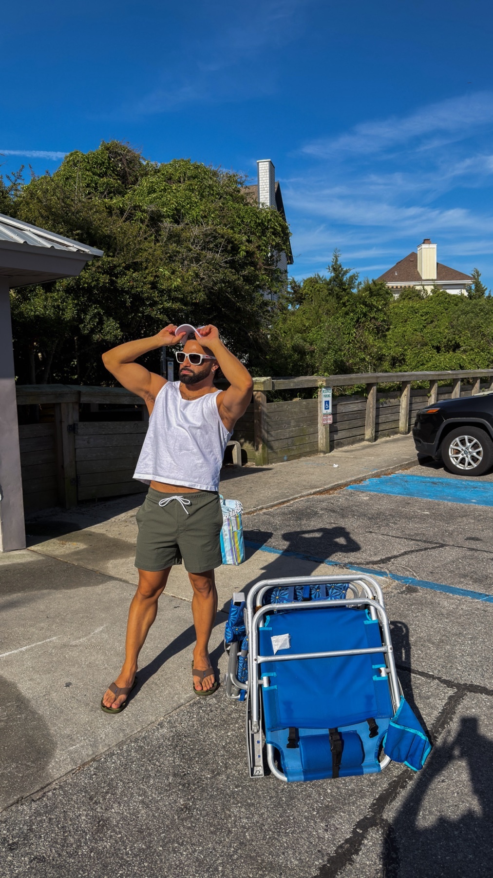 A man poses in a parking lot next to a blue beach chair, wearing sunglasses, a white tank top, olive green shorts, and flip flops. The sky is bright blue with scattered clouds, and lush green trees are visible in the background.