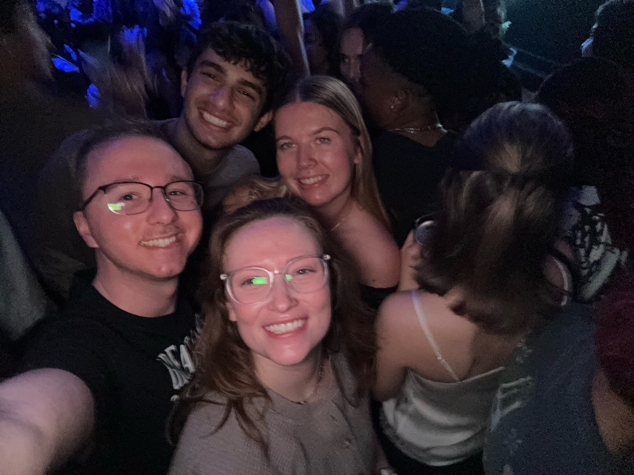 A group of five young adults smile for a selfie at a crowded event, possibly a concert or party, with blue lights illuminating the background.