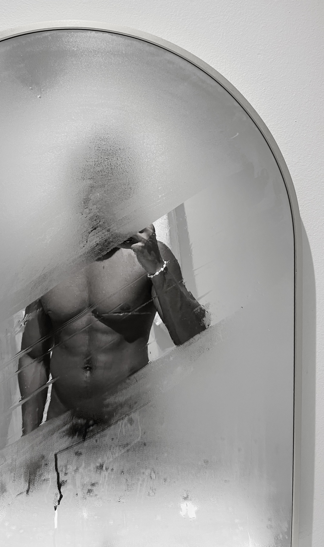 A black and white mirror selfie shows a muscular man with a bare torso reflected in a steamy, arched mirror. The man is wearing jewelry on his wrist and appears to be posing.