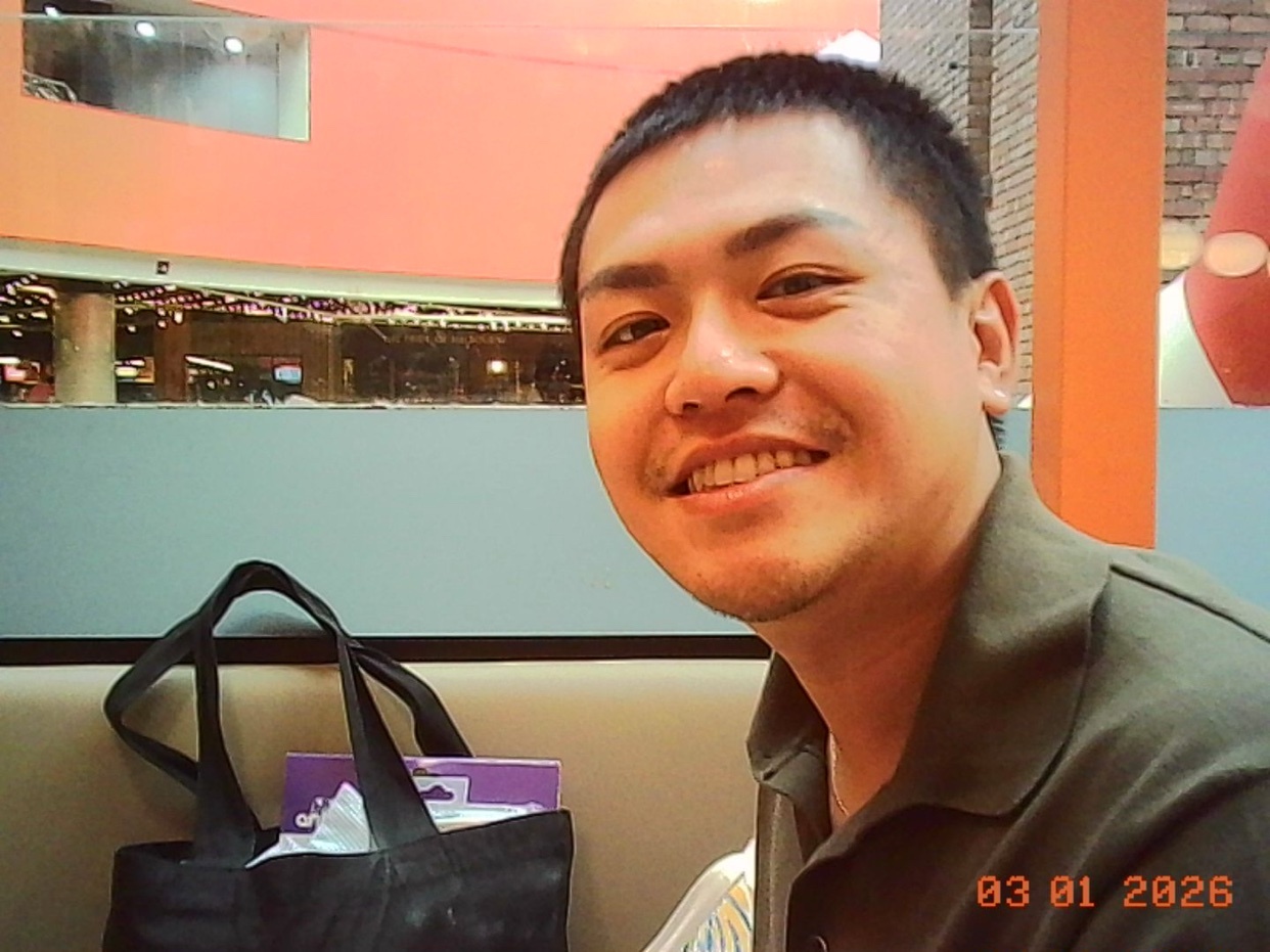 A close-up selfie of an Asian man with short dark hair and a slight smile. He is wearing a dark green collared shirt and is sitting in front of a blurred background that appears to be an indoor shopping mall.