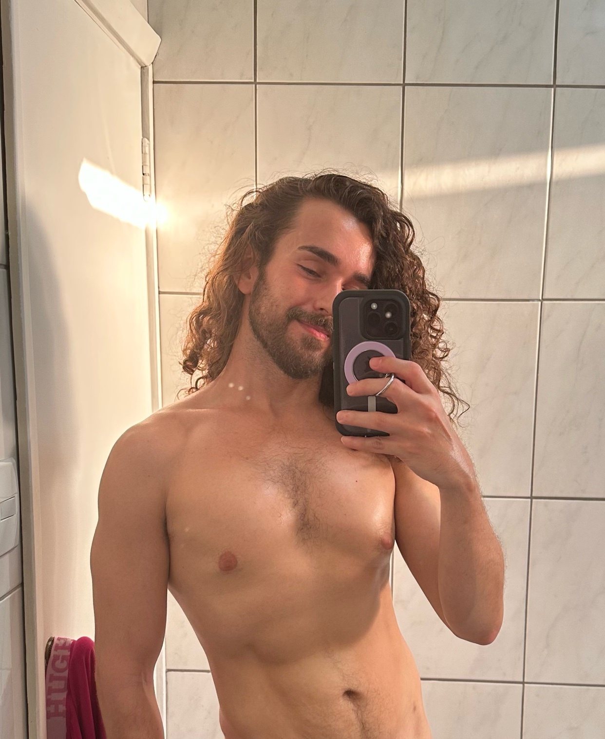 A man takes a mirror selfie, shirtless, in a bathroom. The golden hour light illuminates his curly hair and chest hair.
