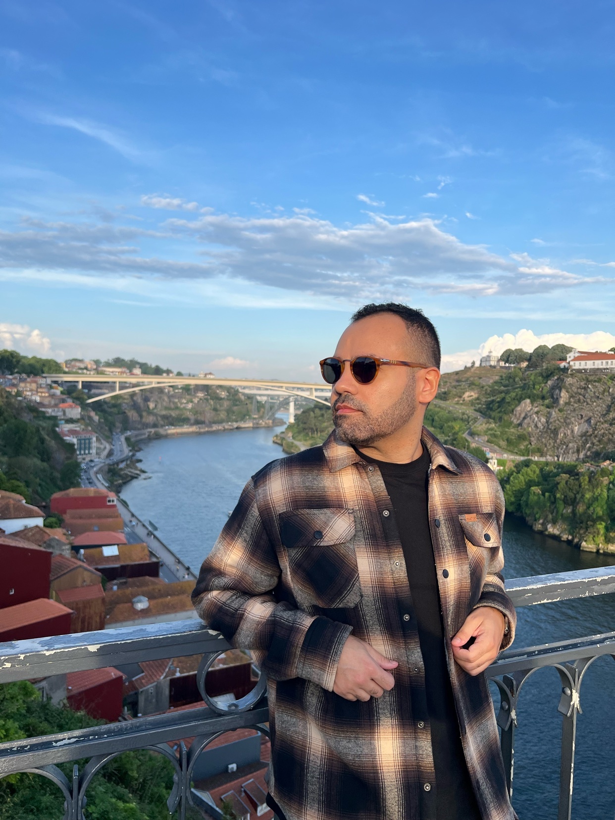A man wearing sunglasses and a plaid shirt poses for a photo on a bridge overlooking a river and a cityscape.