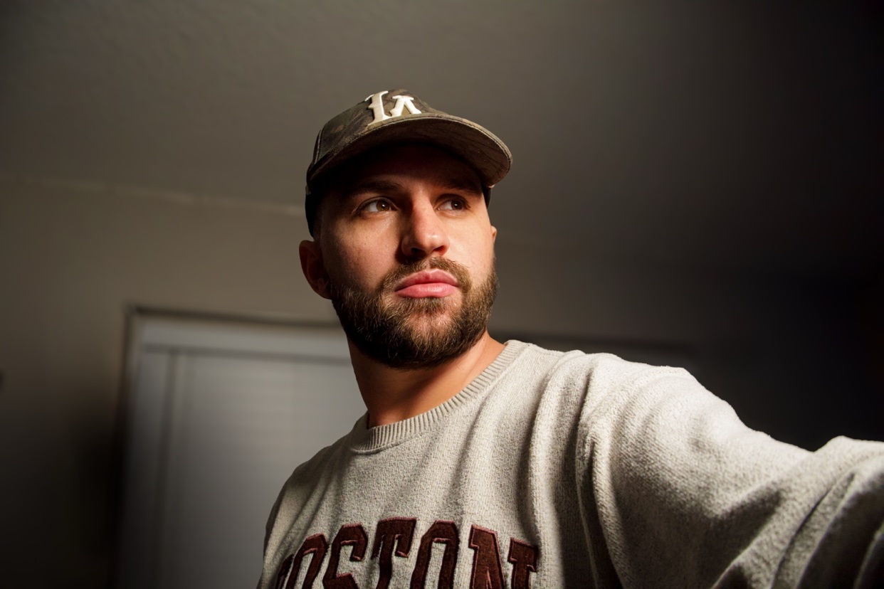 A man with a beard and a baseball cap looks away from the camera. He is wearing a crewneck sweatshirt with the word "Boston" on it.