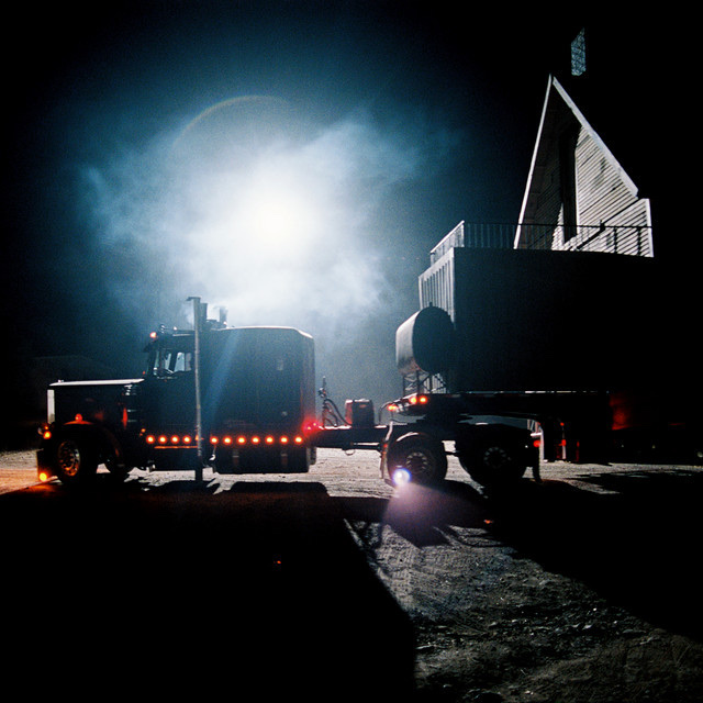 A dark, cinematic shot of two trucks at night. The trucks are silhouetted against a bright light, creating a dramatic and mysterious atmosphere.