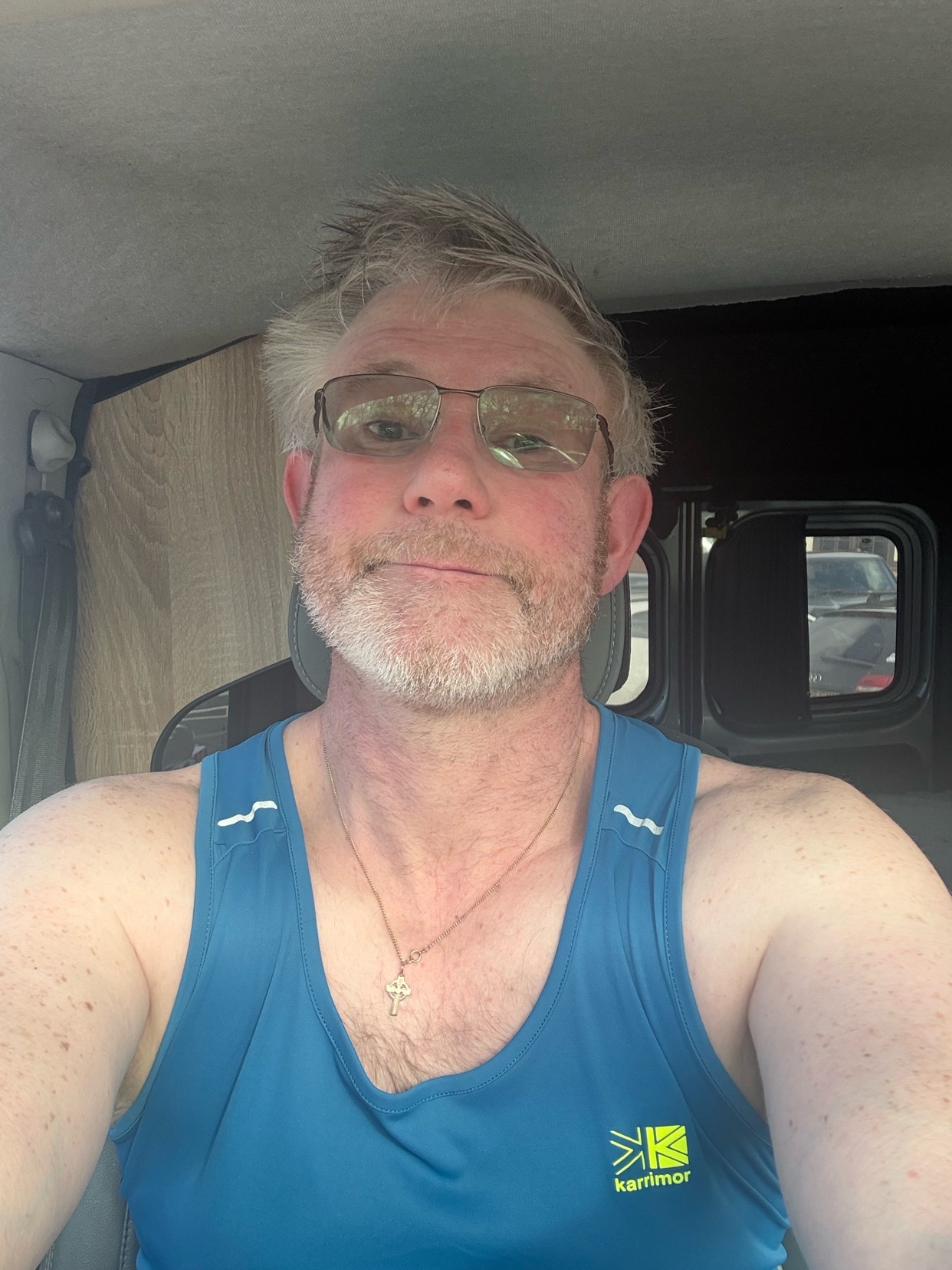 A man with glasses and a beard takes a selfie in a vehicle. He is wearing a blue tank top with a cross necklace.