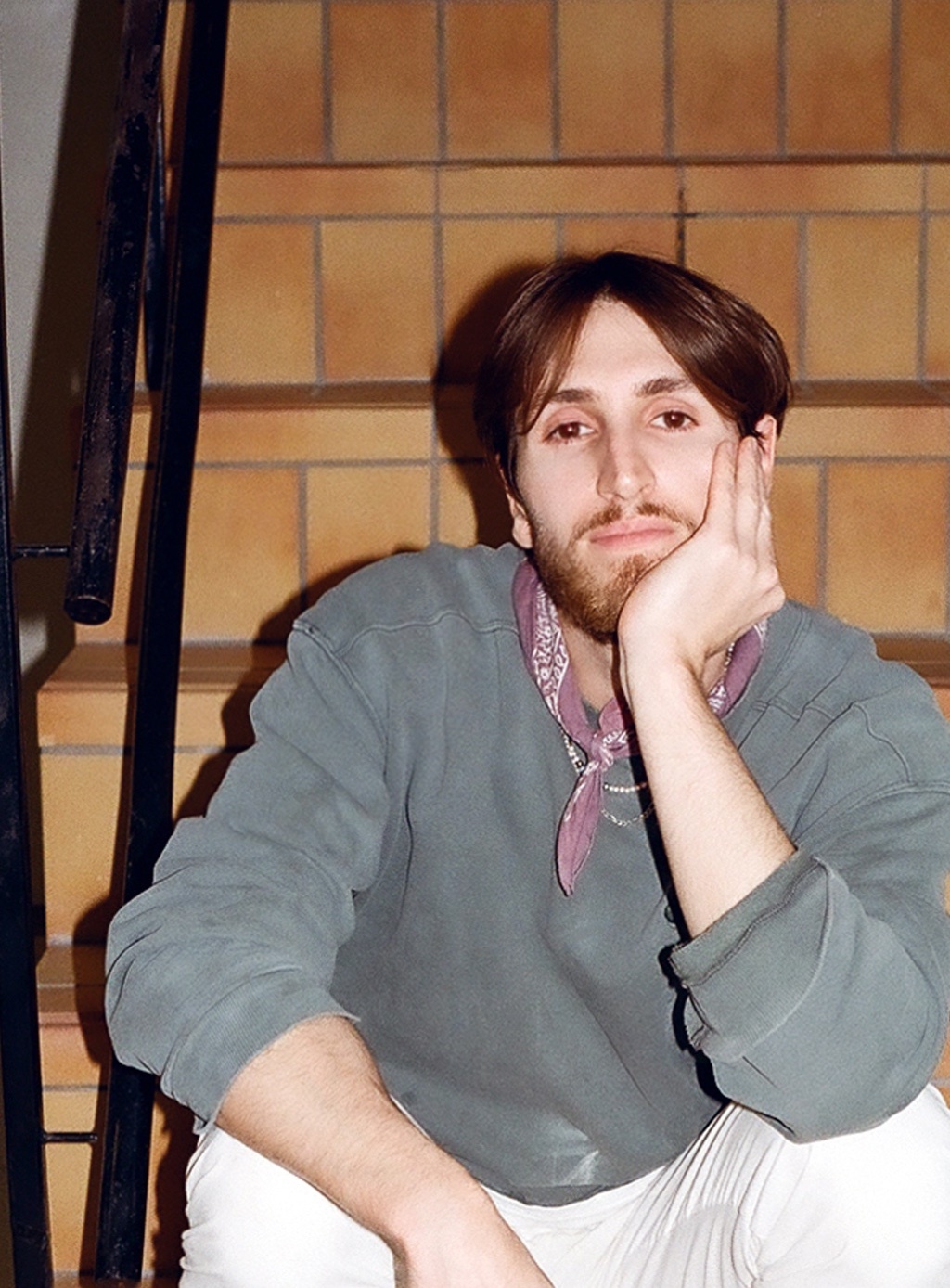 A man poses on a set of stairs, resting his head on his hand. He wears a casual outfit and a bandana.