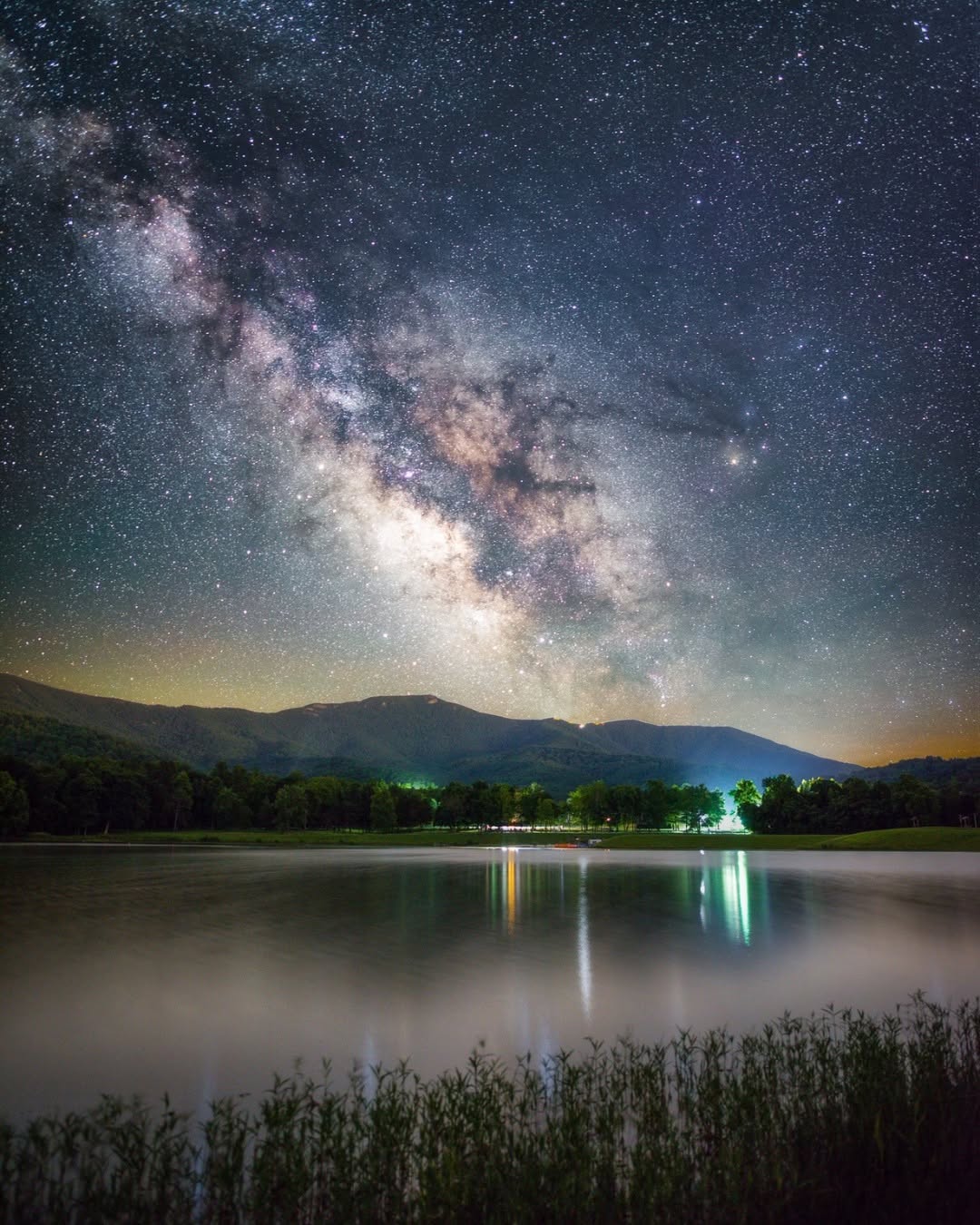 The Milky Way over Lake Arrowhead at night, reflected in the water below the forested ridge