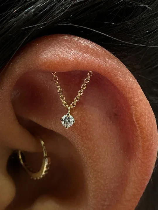 Close-up of a hidden helix ear piercing featuring a gold chain with a dangling solitaire gem end.