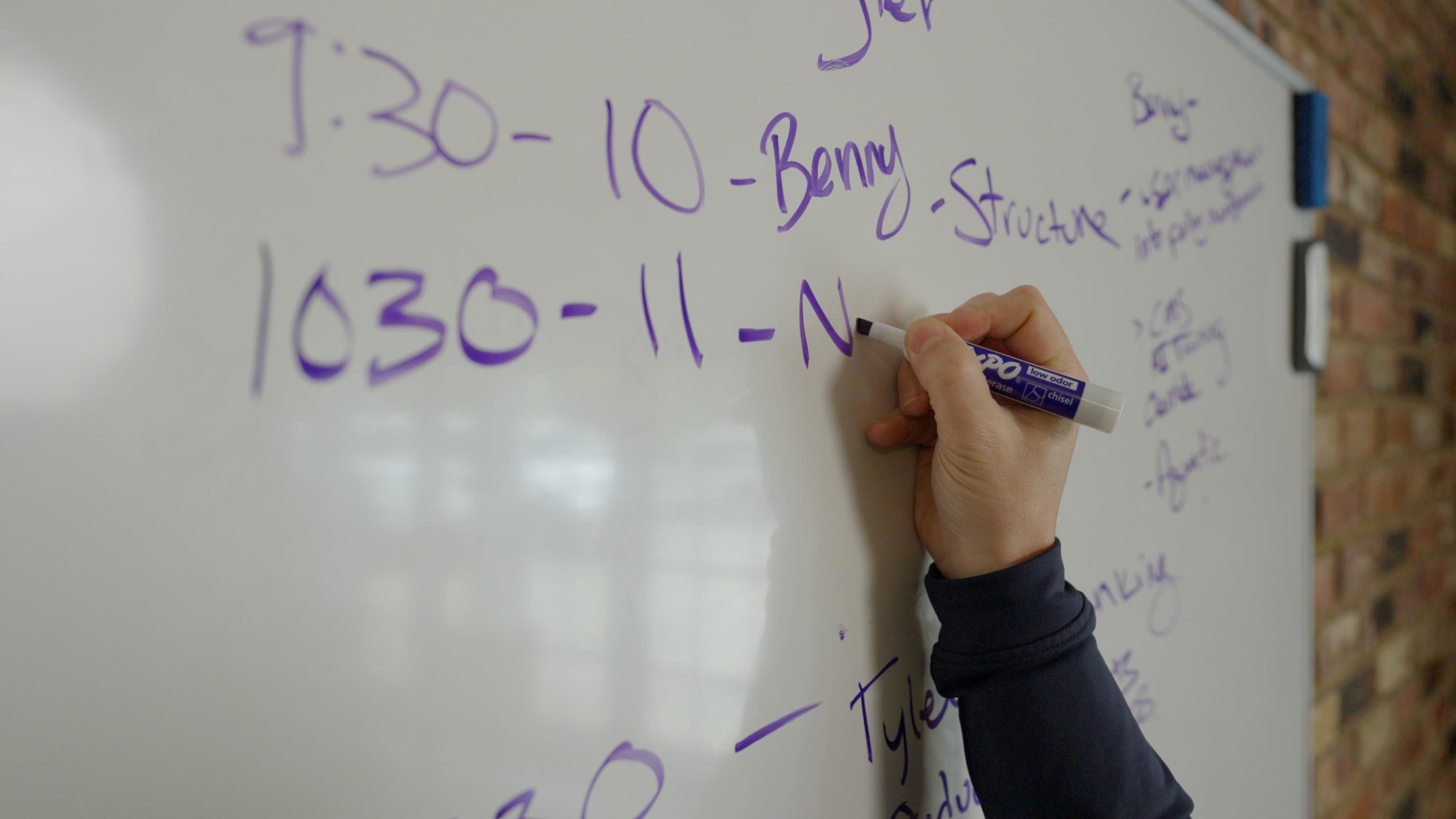 Hand writing on a whiteboard with purple marker, listing schedules and names
