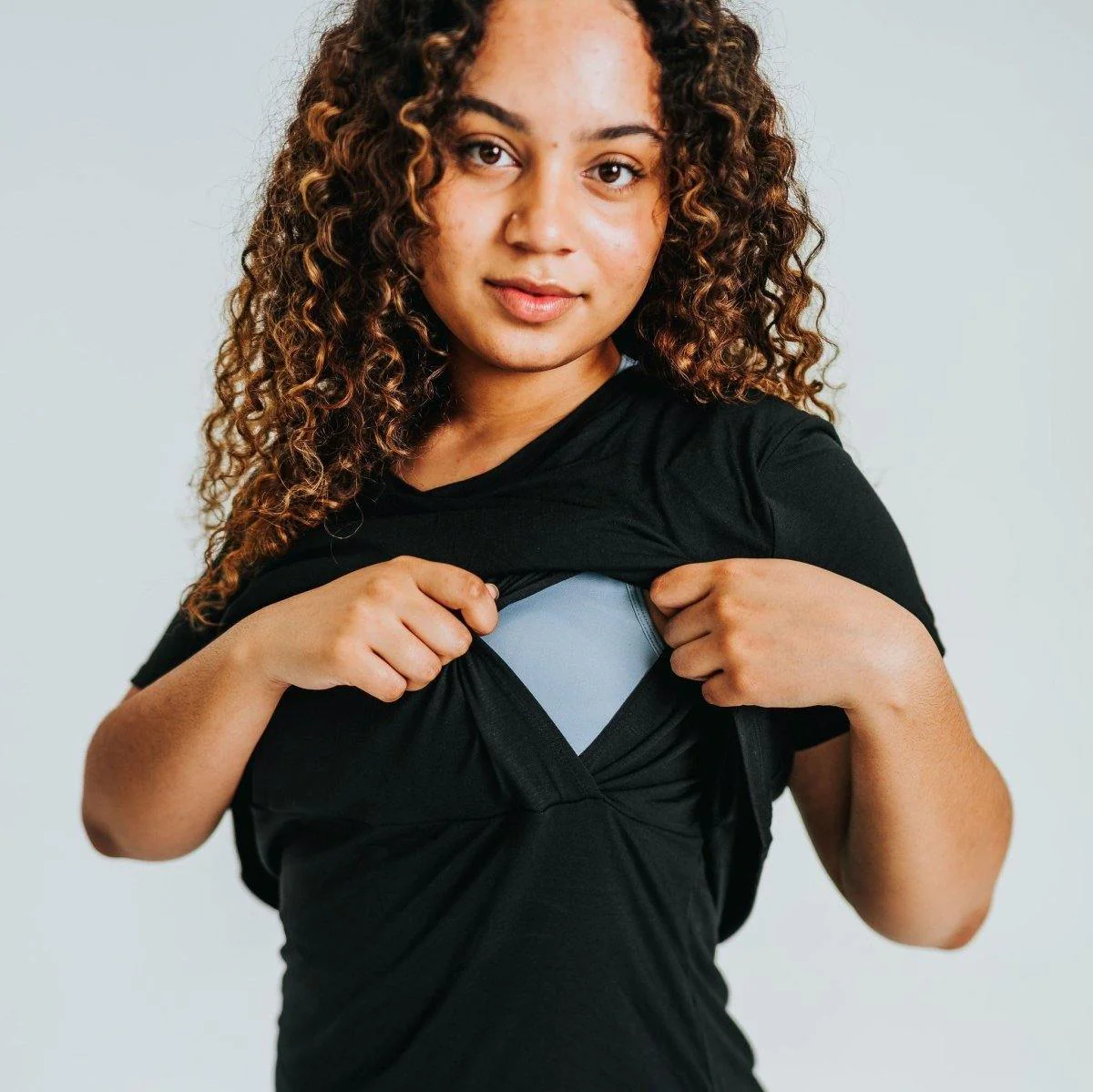 A smiling woman with curly hair demonstrating the dual-layer functionality of a black Senita nursing short-sleeve tee by pulling aside the outer fabric layer to reveal a light blue nursing bra underneath.
