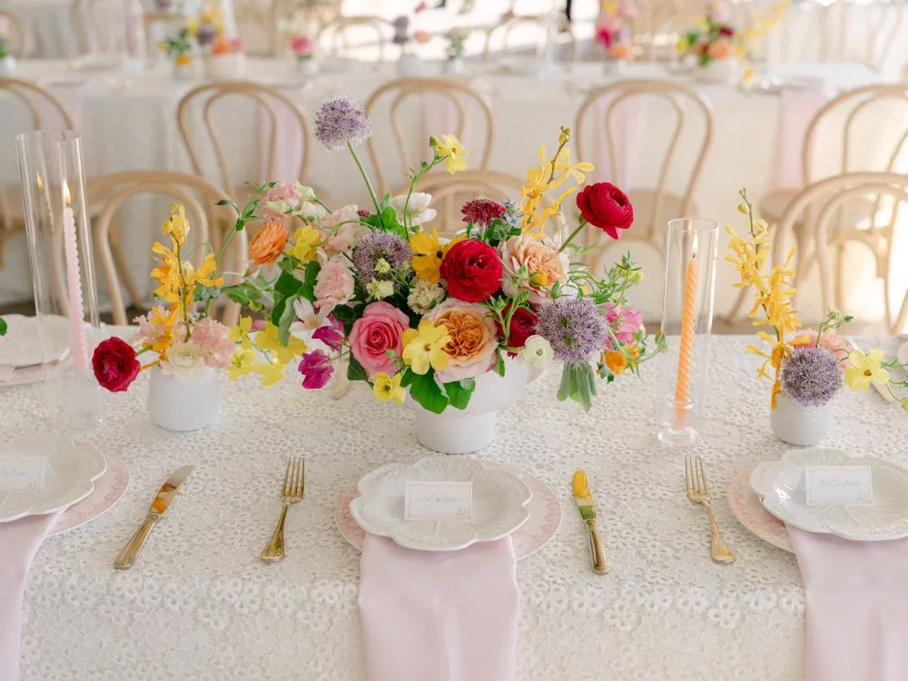 Bright wedding table setting with white textured linen, pastel napkins, colorful flowers, and gold flatware.