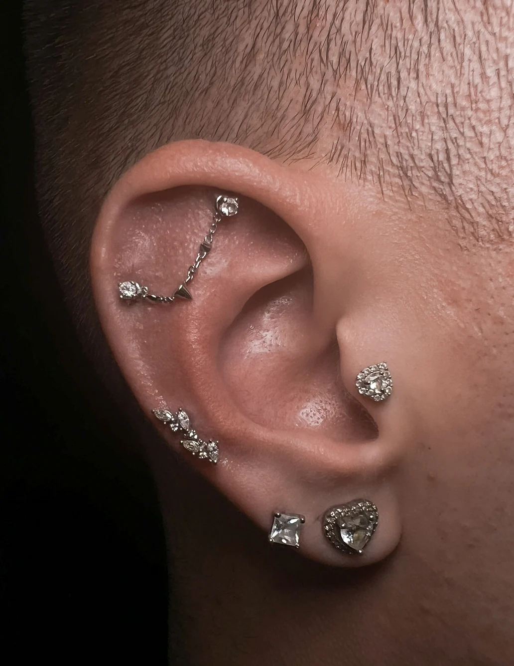 Close-up of an ear featuring a cohesive silver and diamond curation. A thin silver chain connects two piercings in the flat/helix area. The set is complemented by a heart-shaped diamond stud in the tragus, a marquise-cut crystal cluster in the mid-helix, and heart-shaped silver earrings in the lobe.