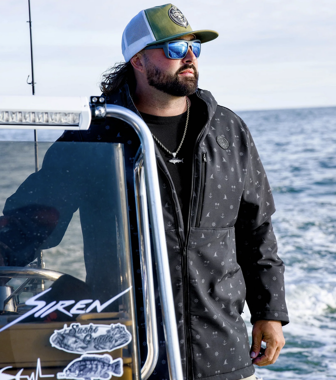A bearded man wearing a black Anchor Jacket with a nautical pattern, a green and white trucker hat featuring a Dixxon crest, and blue mirrored sunglasses. He is standing on a boat titled "Siren," embodying the adventurous maritime lifestyle the jacket is built for.