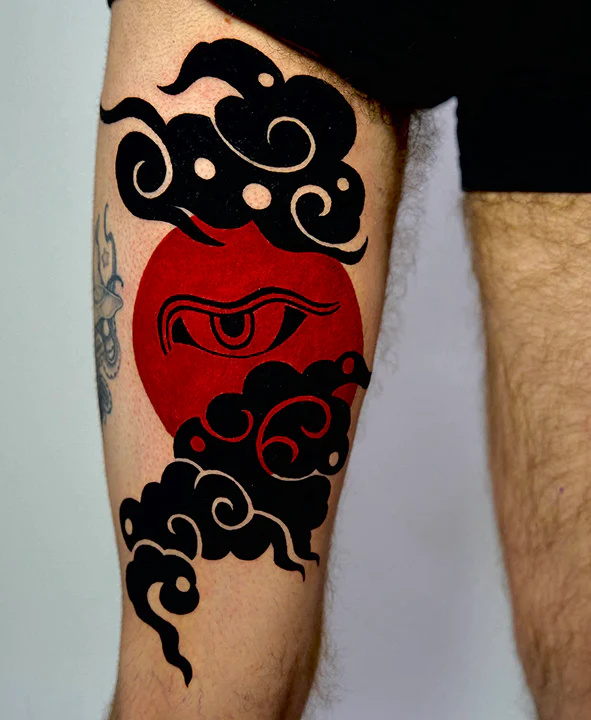 A striking neo-traditional tattoo on a person's thigh featuring a bold red circle containing a stylized eye. The eye is surrounded by thick, solid black clouds with white accents, creating a high-contrast and graphic appearance.