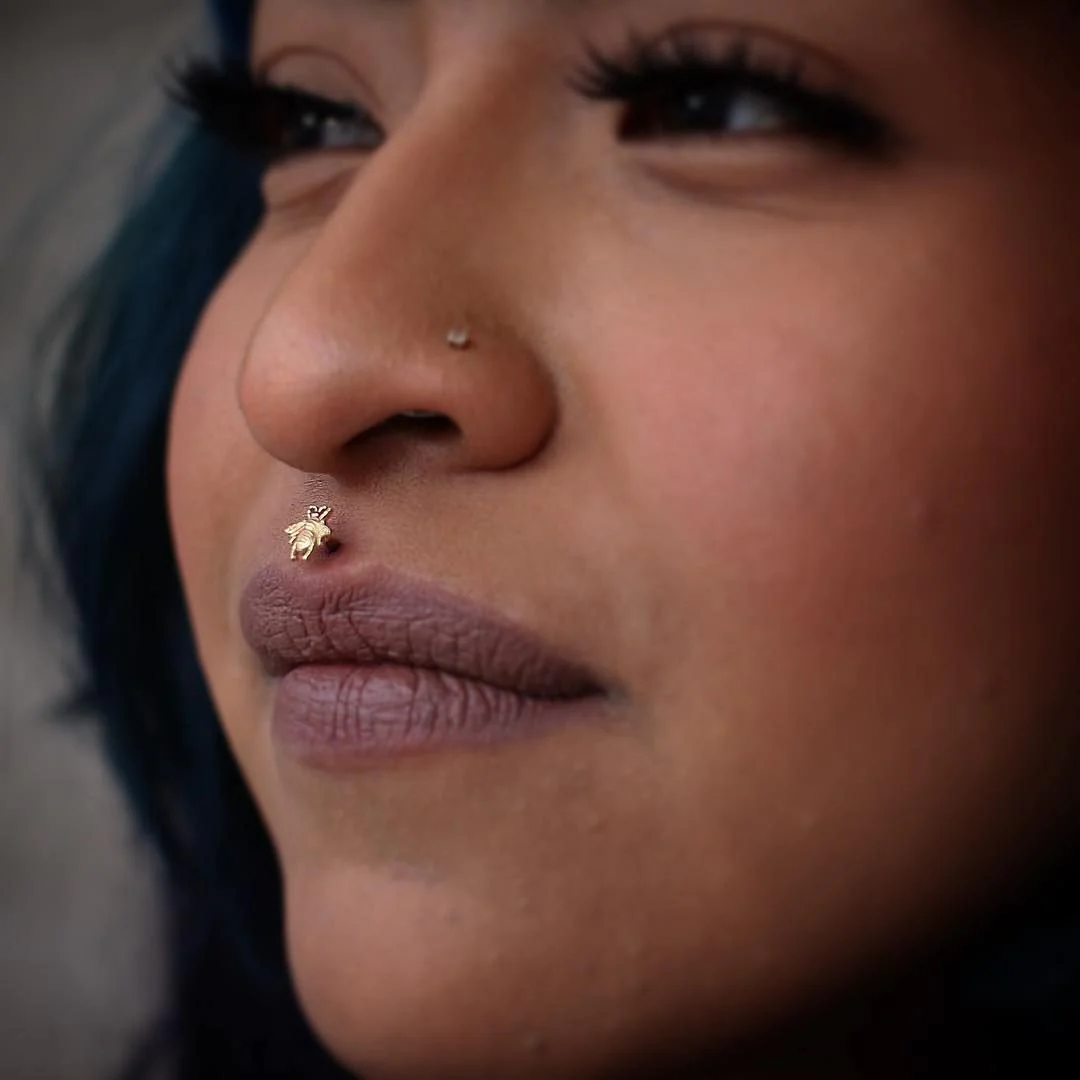 Profile view of a person with blue hair showcasing a gold philtrum piercing featuring a highly detailed, small honeybee stud sitting perfectly above the upper lip.