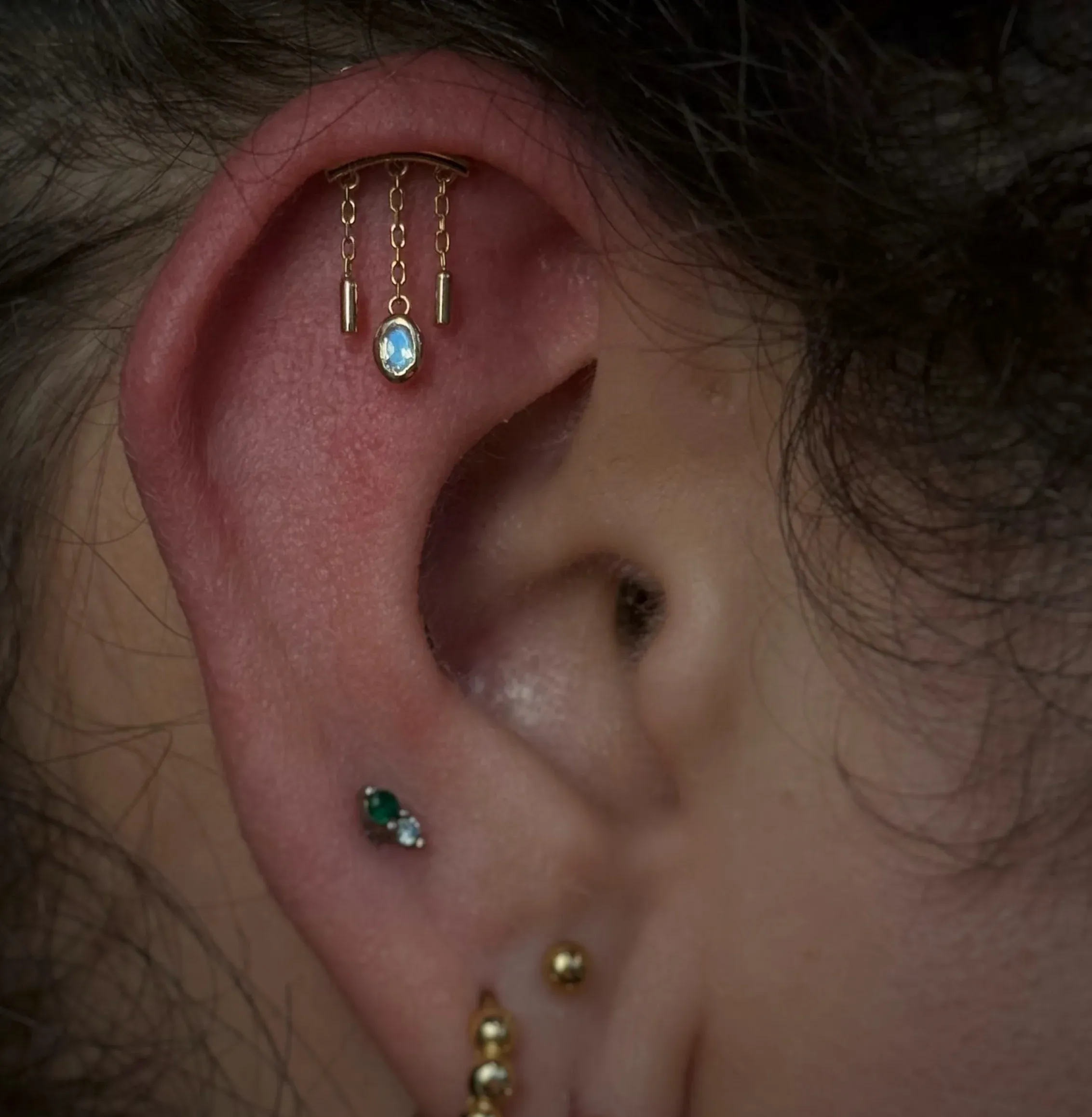 A flat ear piercing with a gold chandelier charm holding dangling chains and a center gem.