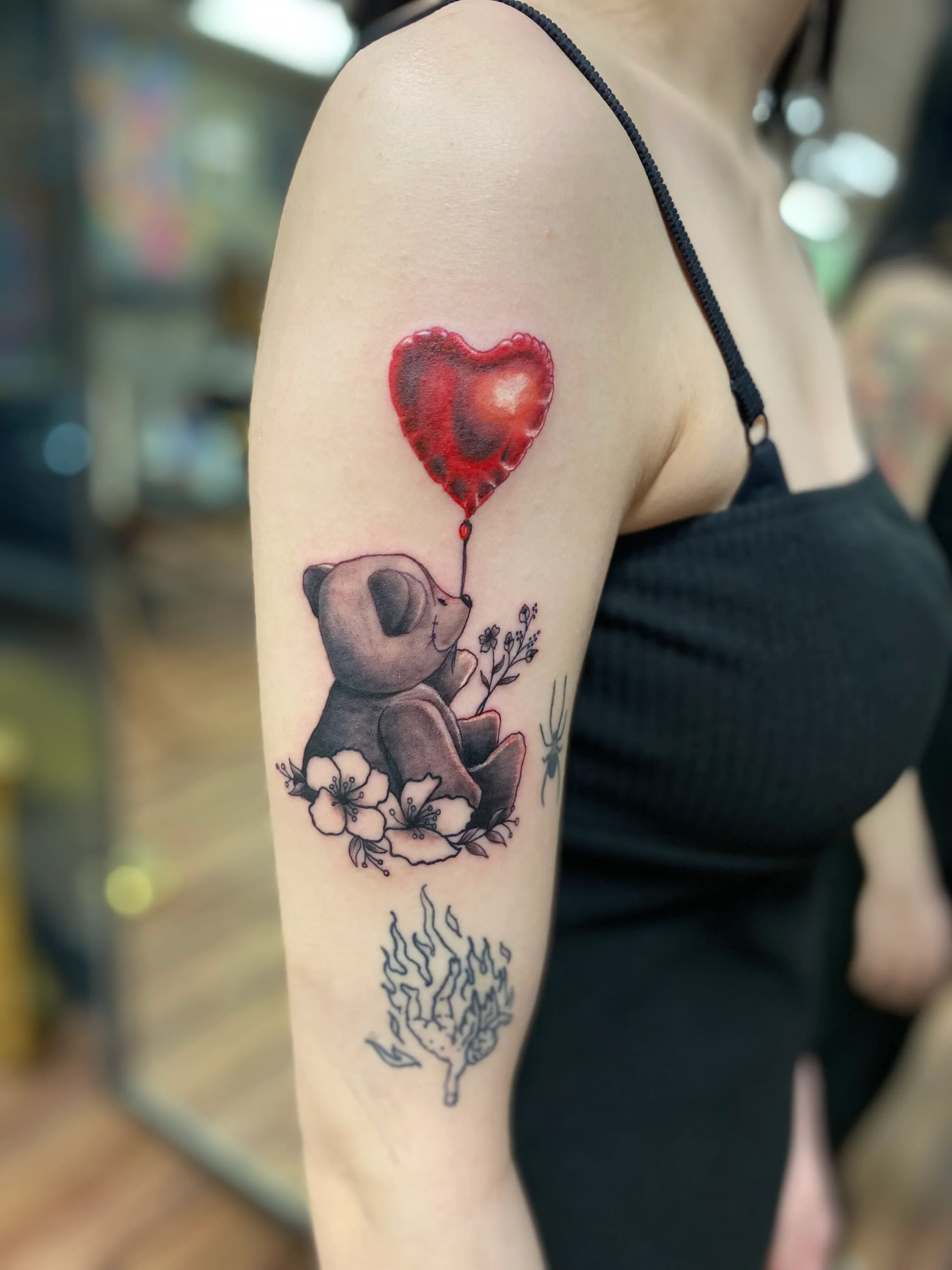 Upper arm tattoo of a small grey teddy bear holding a red heart balloon with floral details underneath.