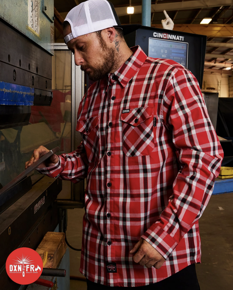 A bearded man in a white mesh-back cap operating a Cincinnati industrial press brake. He is wearing a red, white, and black Dixxon flame-resistant (FR) flannel, emphasizing the brand's commitment to high-quality gear for the working class.