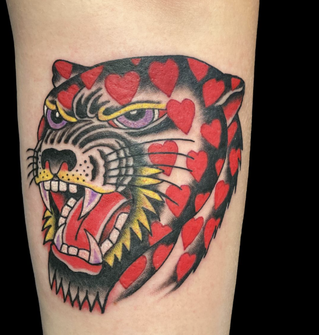 Close-up of a bold, traditional style panther head tattoo on an arm, its black fur dotted with a pattern of vibrant red hearts. The panther has an open mouth showing large fangs and an purple-tinted, intense expression.