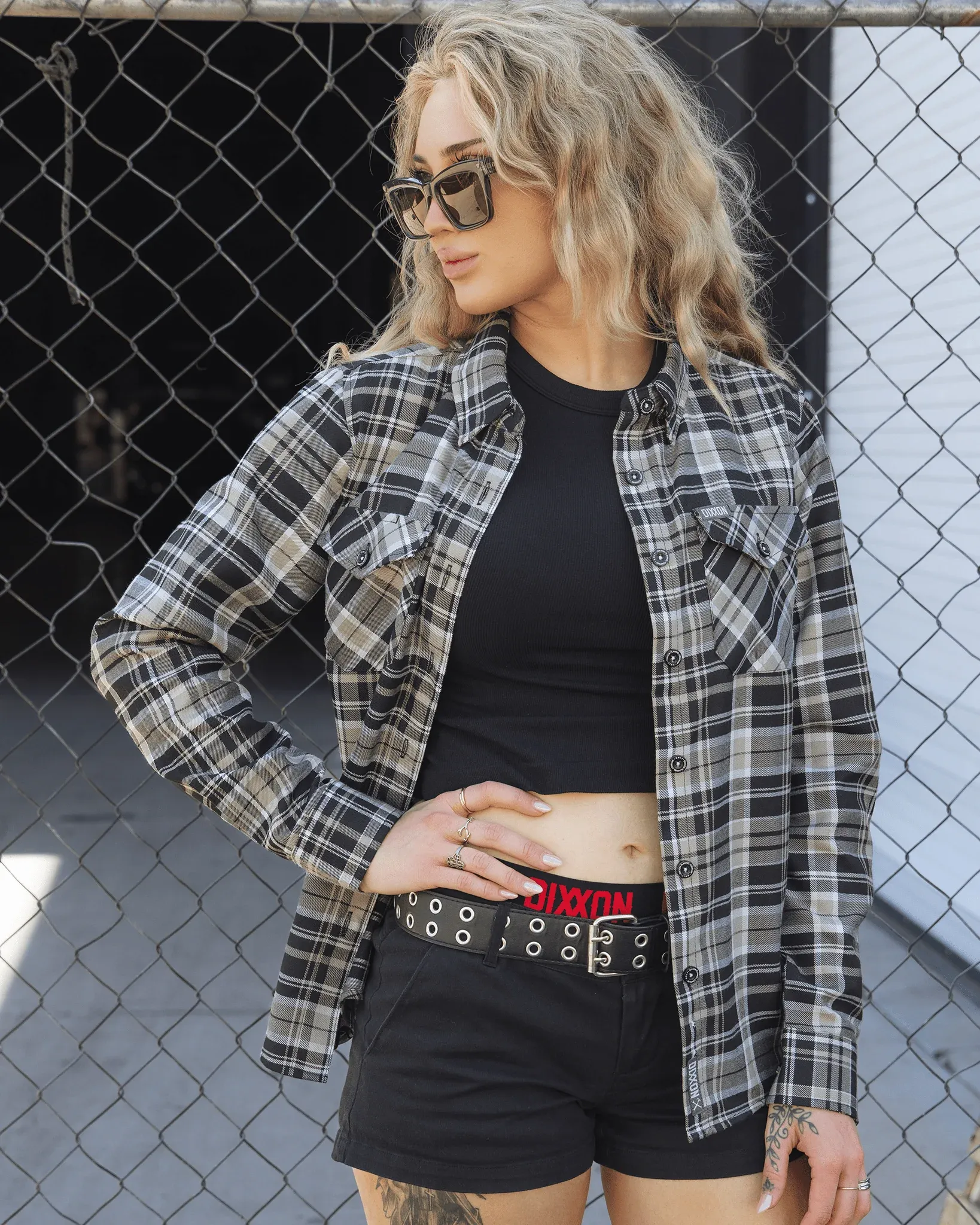 Blonde woman wearing an unbuttoned black and grey Dixxon plaid flannel shirt over a black crop top.