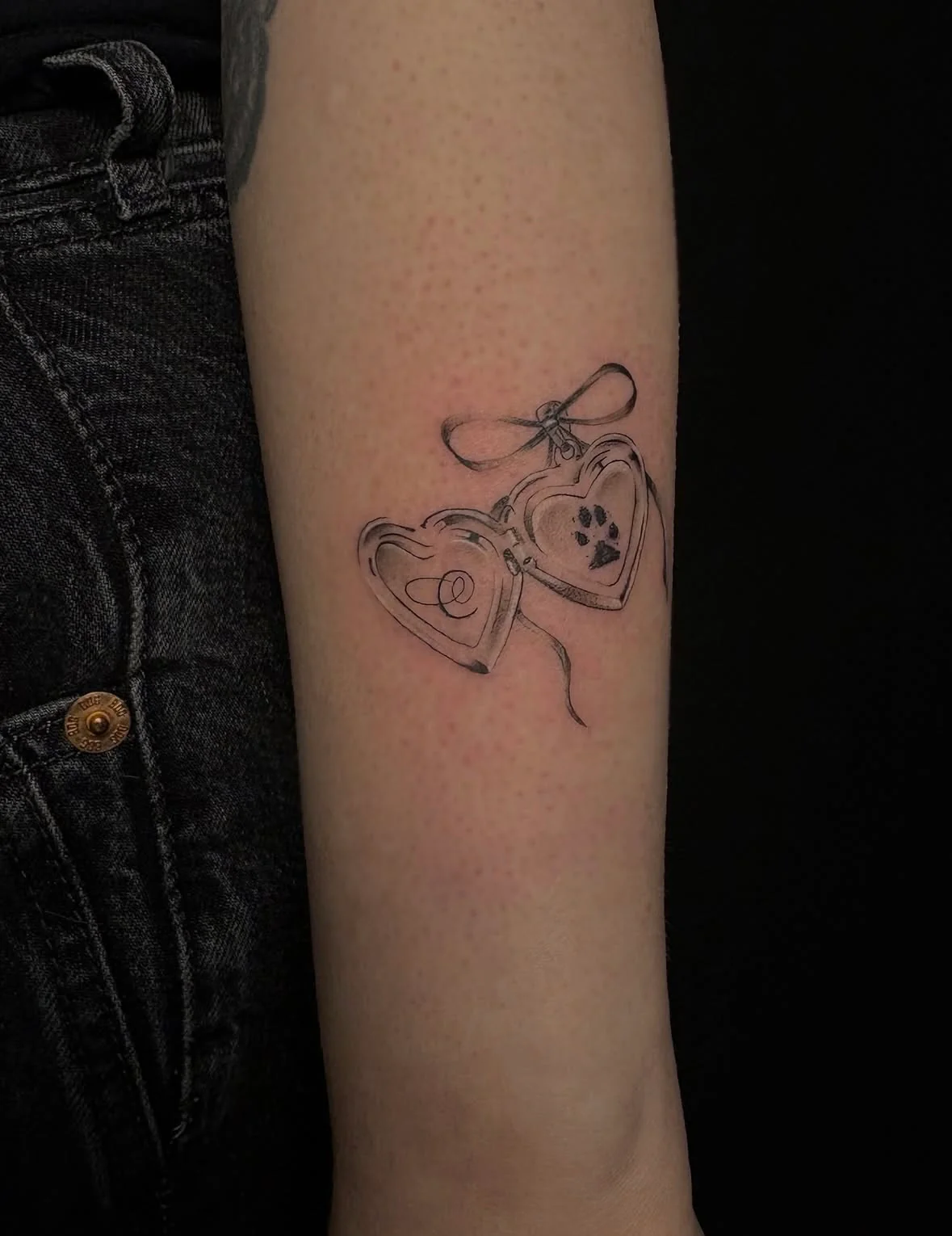Delicate fine-line tattoo on an arm featuring an open, heart-shaped locket tied with a dainty bow. The left side of the locket displays a minimalist cursive initial, while the right side holds a small black dog paw print.