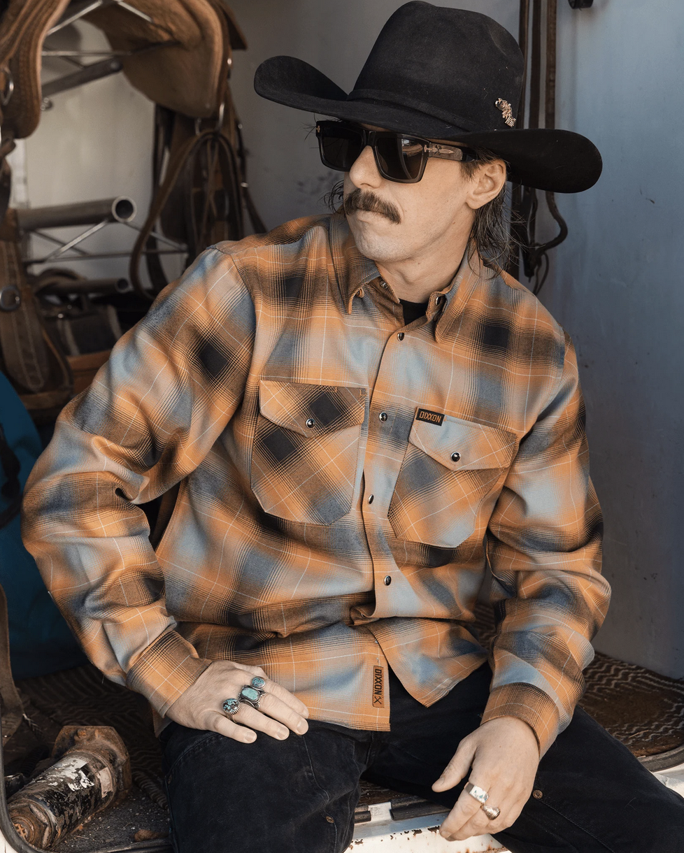 A man with a mustache wearing a black cowboy hat and a Dixxon flannel shirt with a tan, blue, and black plaid pattern. He is seated in a rugged, sunlit environment, showcasing the flannel's mitered edge pockets and reinforced construction.