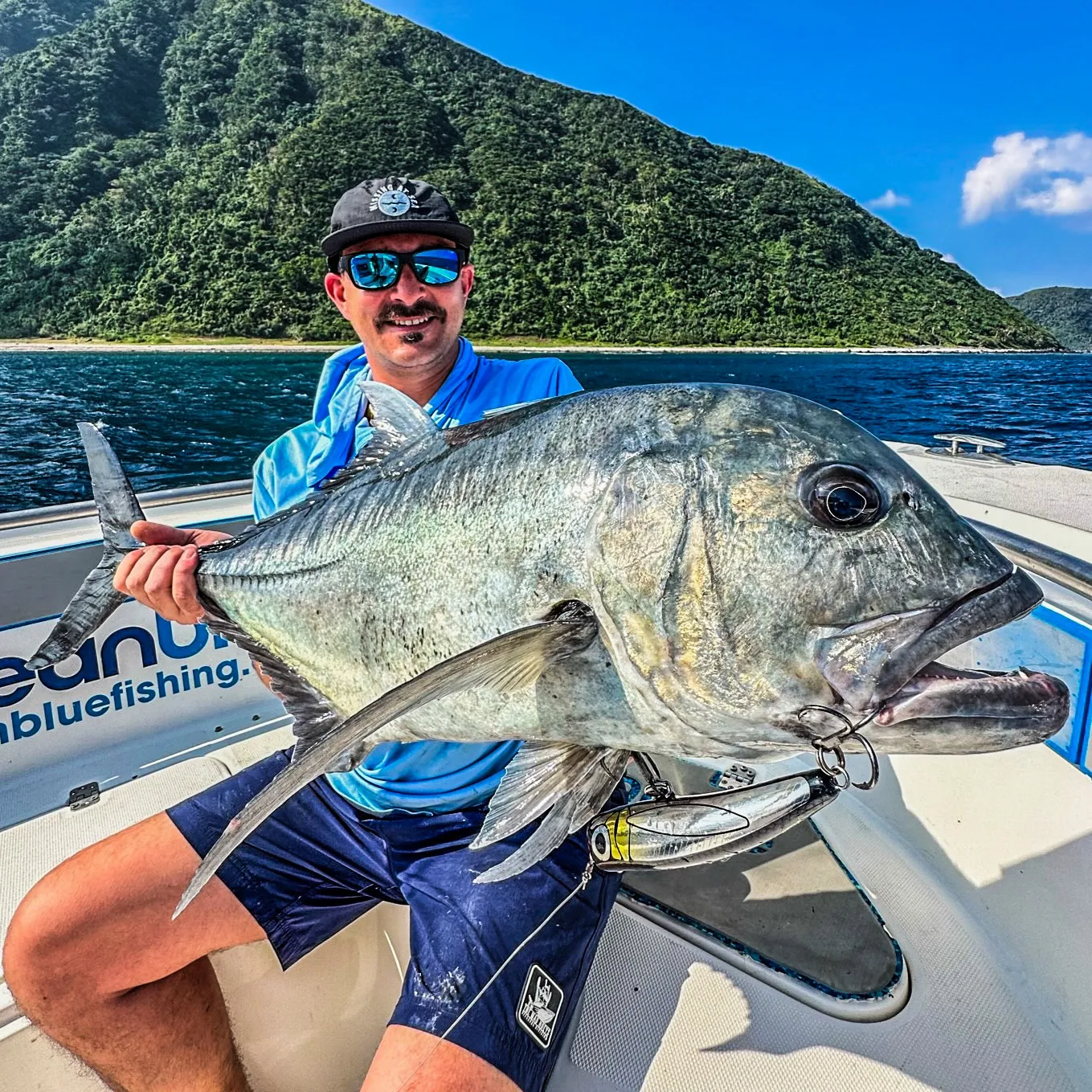 Angler holding a large giant trevally (GT) on an Ocean Blue Fishing Adventures boat in Vanuatu, with a lush tropical island coastline behind.
