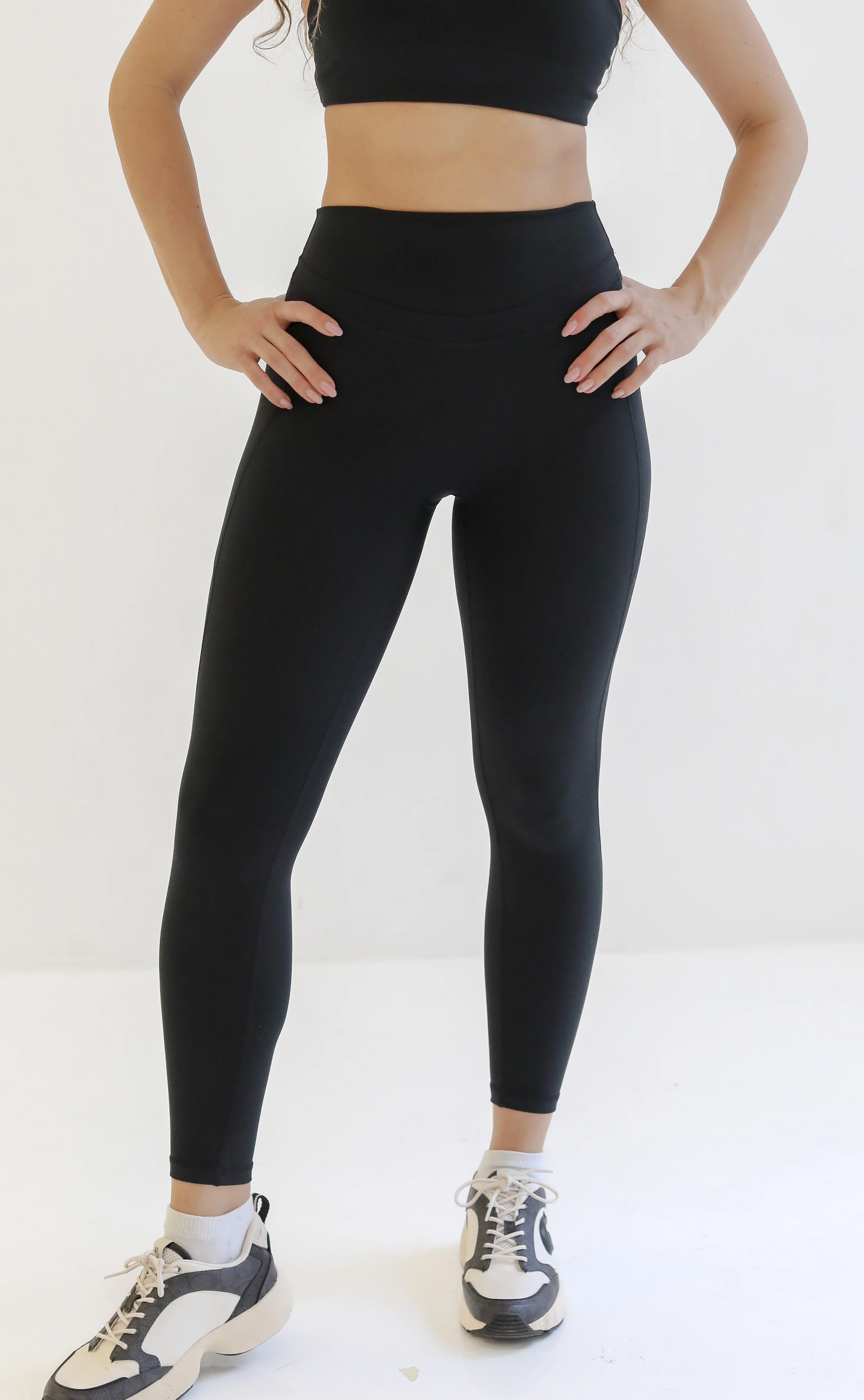 Front view of a woman standing in full-length high-waisted black athletic leggings paired with a matching black sports bra and white sneakers.