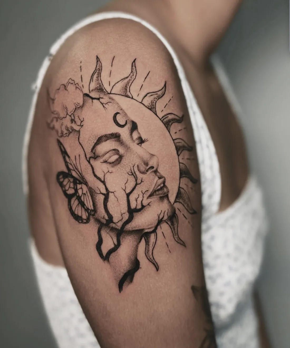 Black and grey upper arm tattoo featuring a woman's face split into a sun, moon crescent, and butterfly design.