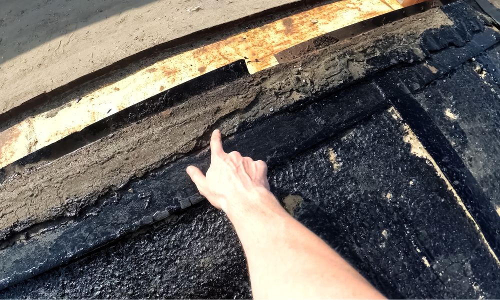 A person's hand pointing to a damaged and deteriorated area of old asphalt roofing material near the exposed wood underlayment where roof flashing needs to be repaired.