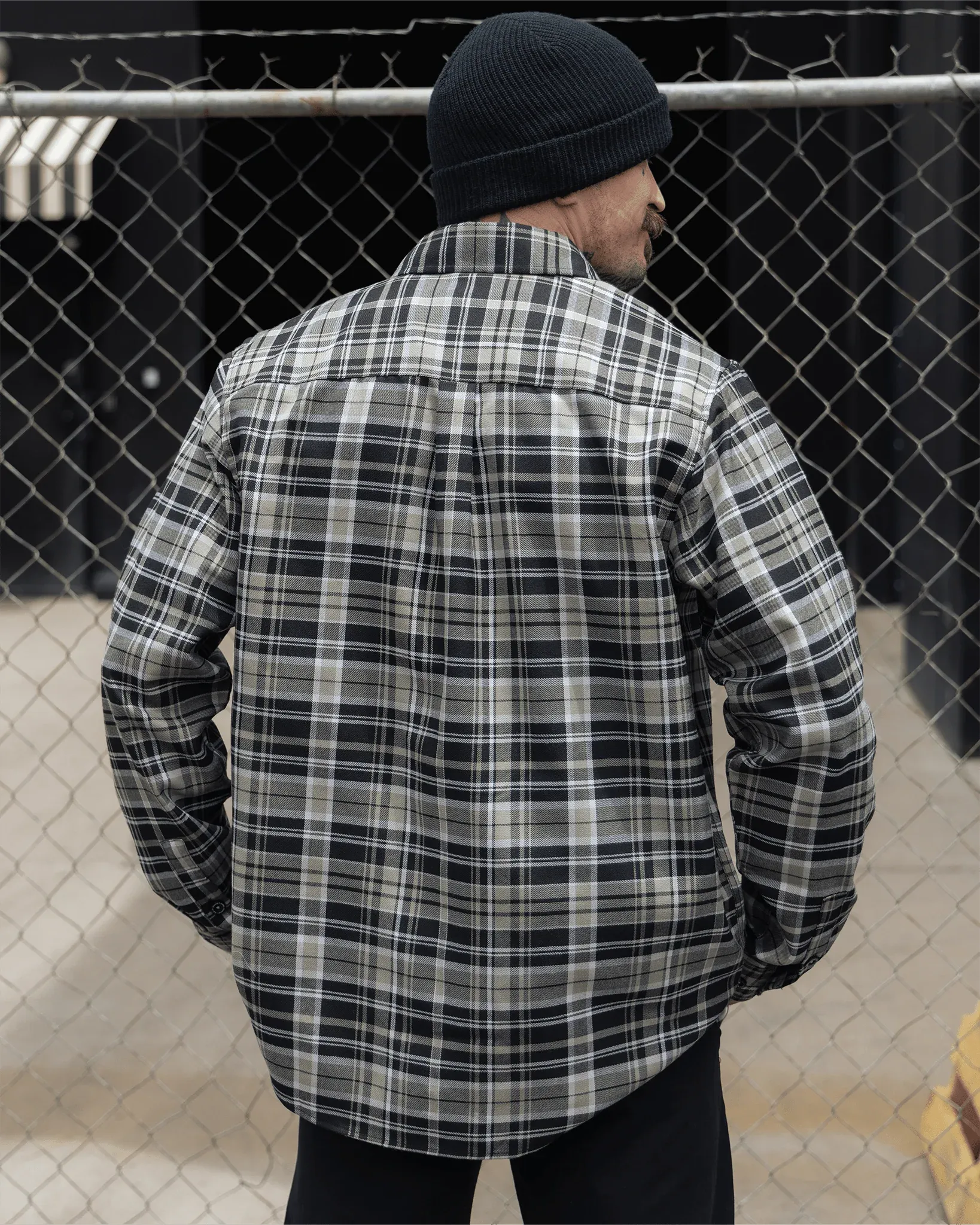 Back view of a guy in a beanie wearing the black and white Jinx plaid flannel by a chain-link fence.