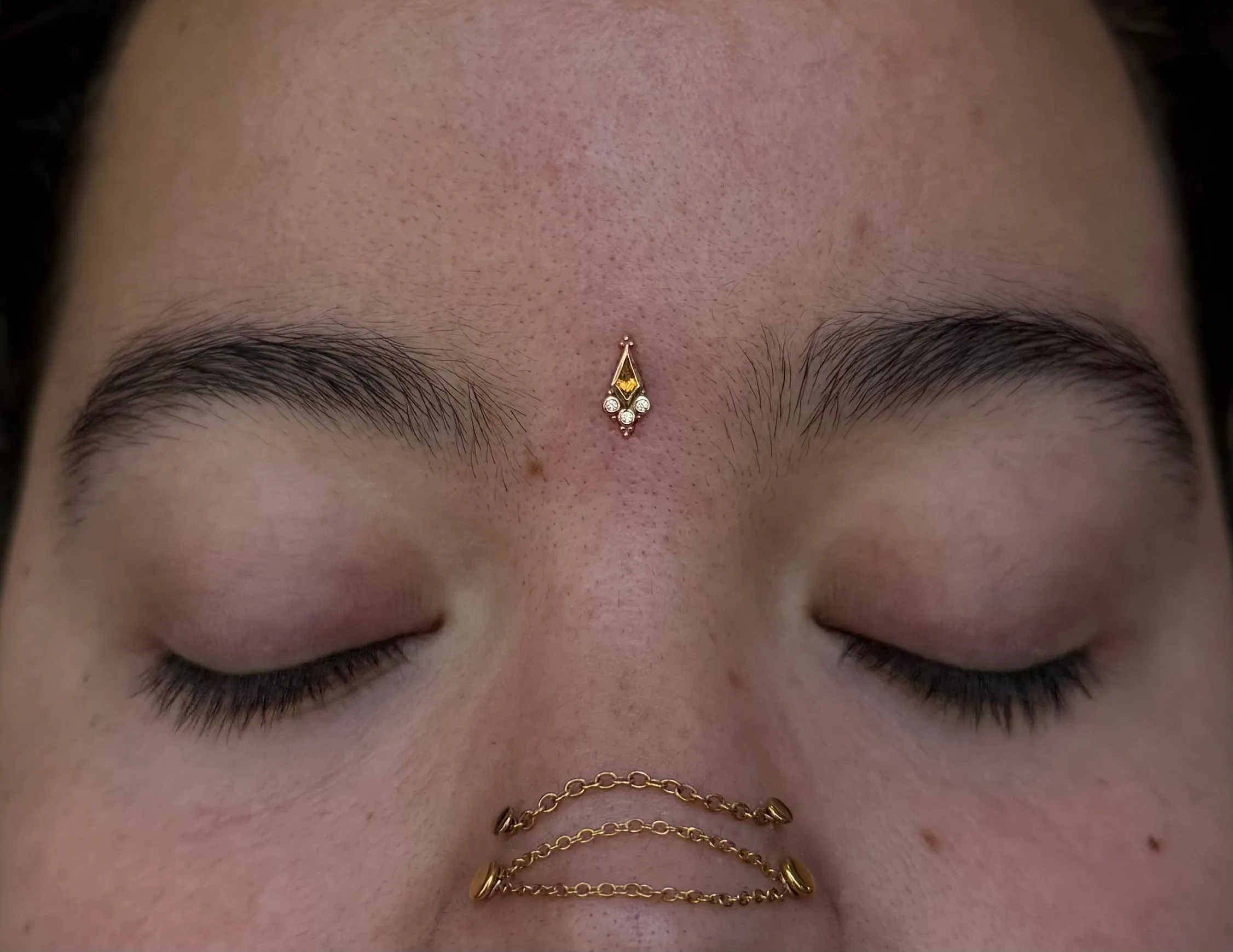 A bindi dermal anchor piercing between the eyebrows paired with gold nose chain jewelry.