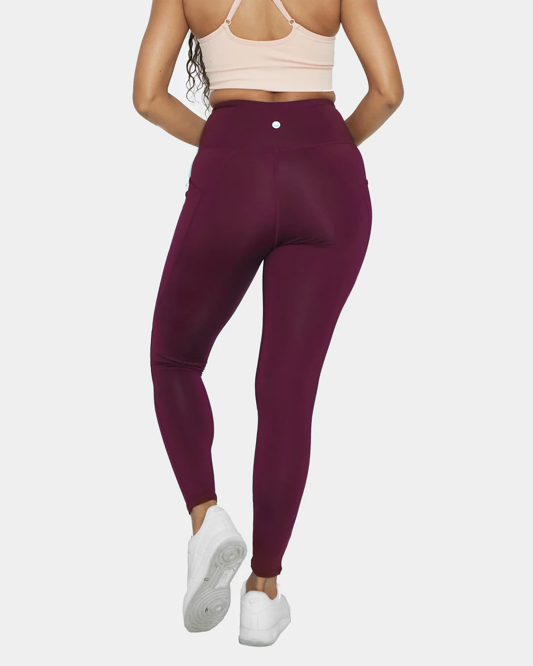 Rear view of a woman showcasing high-waisted mulberry purple athletic leggings with subtle seam details and a small circular logo on the back waistband, paired with white sneakers and a light pink sports bra.