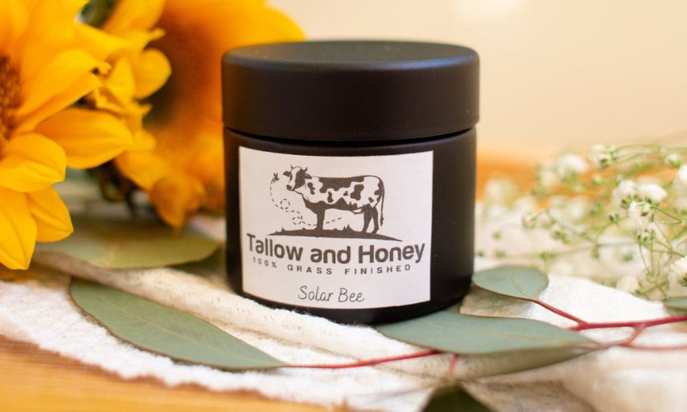 A small black jar of Tallow and Honey "Solar Bee" 100% grass-finished best tallow sunscreen, resting on a white fabric surrounded by sunflowers, eucalyptus leaves, and baby's breath.