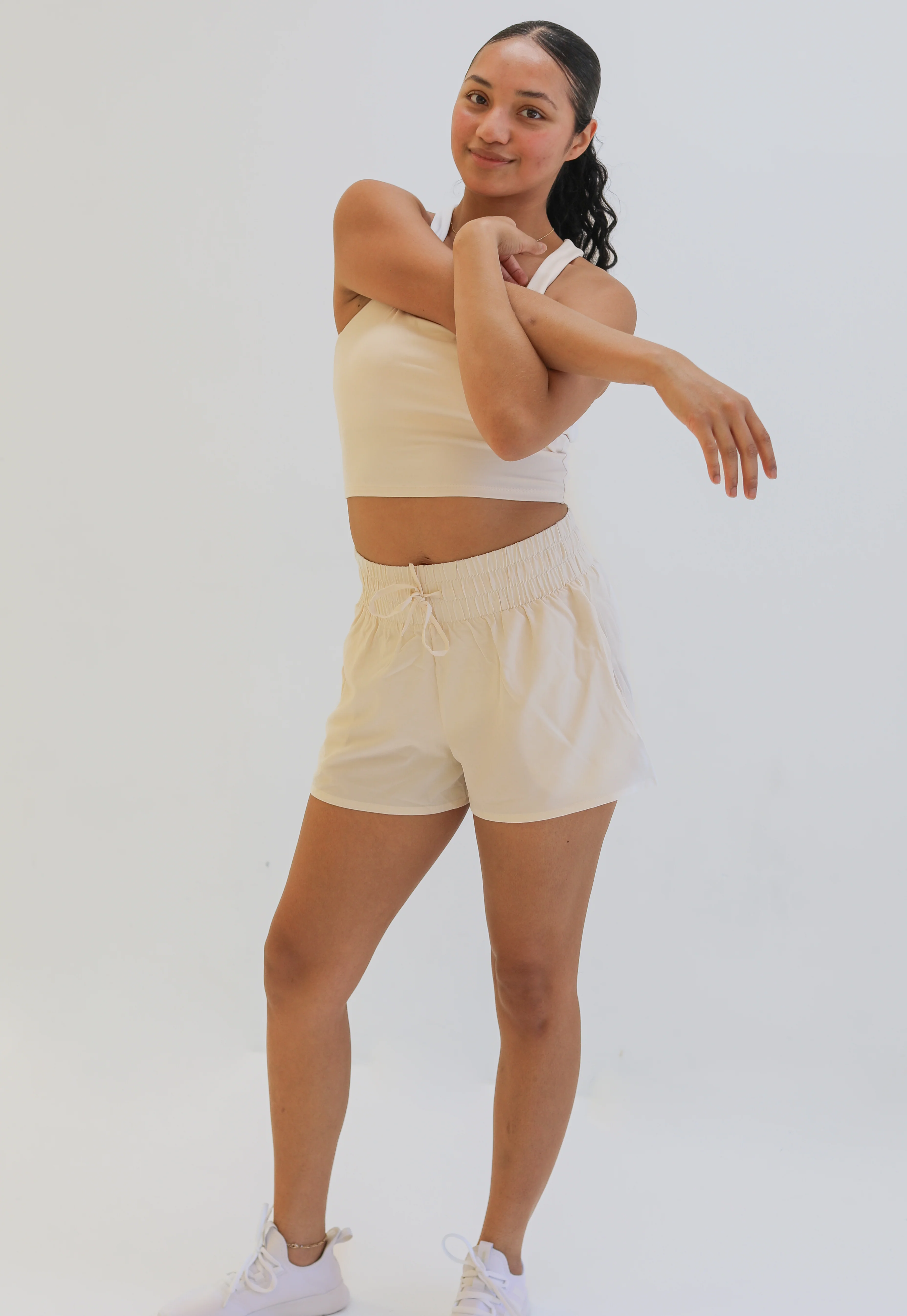 Full-body studio shot of a woman stretching her arms while wearing a cream-colored cropped tank top and matching loose-fitting, cream woven exercise shorts with a wide gathered elastic waistband.