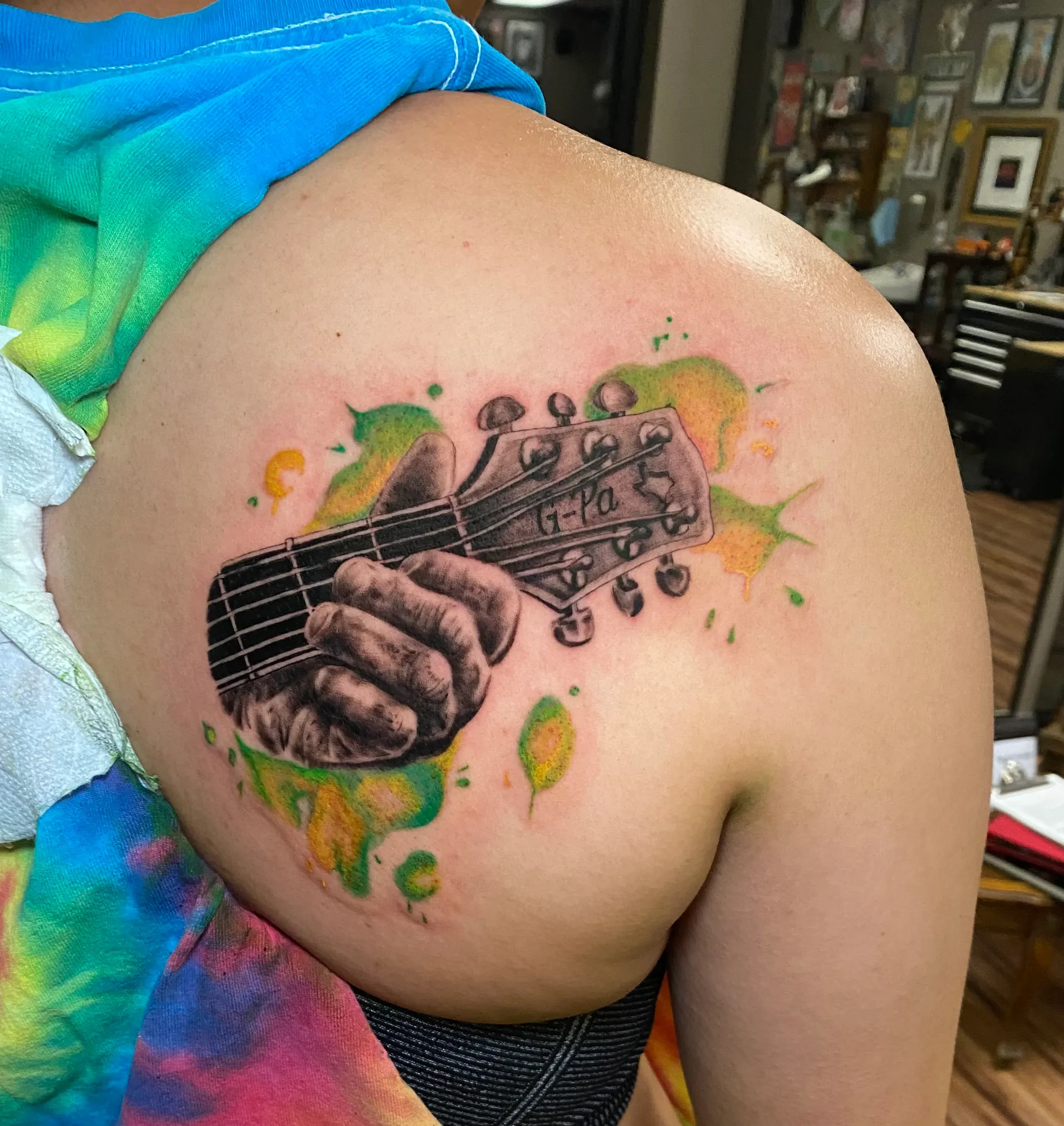 Black and grey hand playing a guitar headstock tattoo surrounded by green and yellow watercolor splashes on a shoulder.