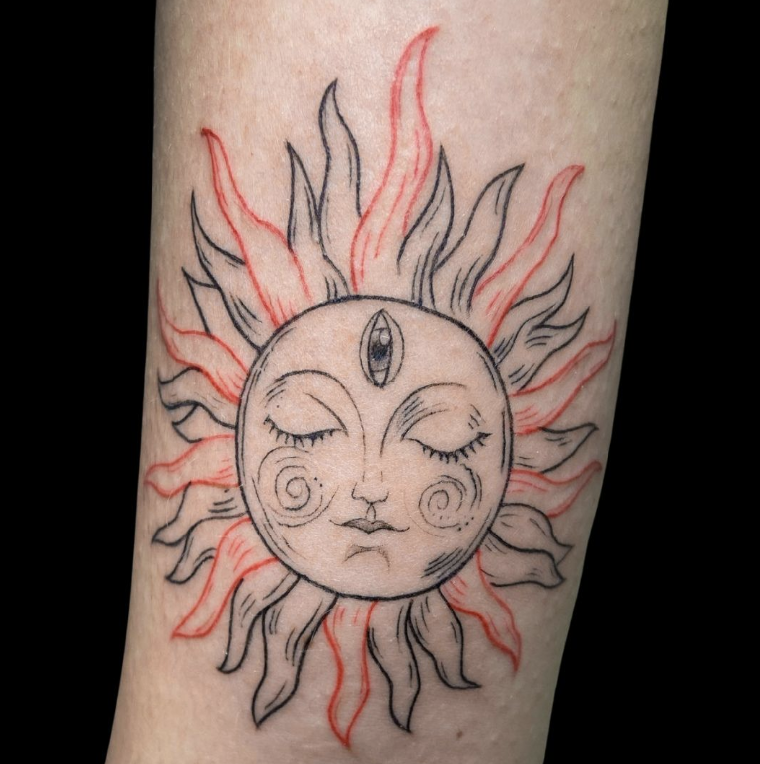 A hand with light skin shows a celestial sun tattoo in red and black ink. The sun features a face with closed eyes, rosy swirling cheeks, and a stylized third eye in its forehead.