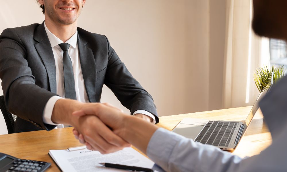 A professional real estate agent in a suit shaking hands with a client over a desk with a contract, successfully finalizing a luxury home transaction.